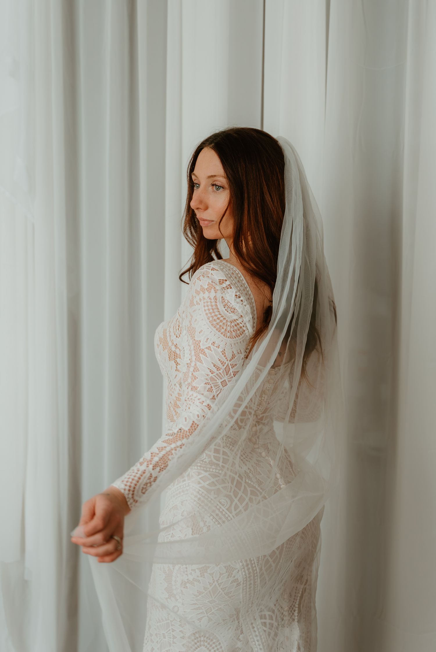 A bride in a long-sleeve lace wedding gown and veil stands gracefully beside sheer white curtains.