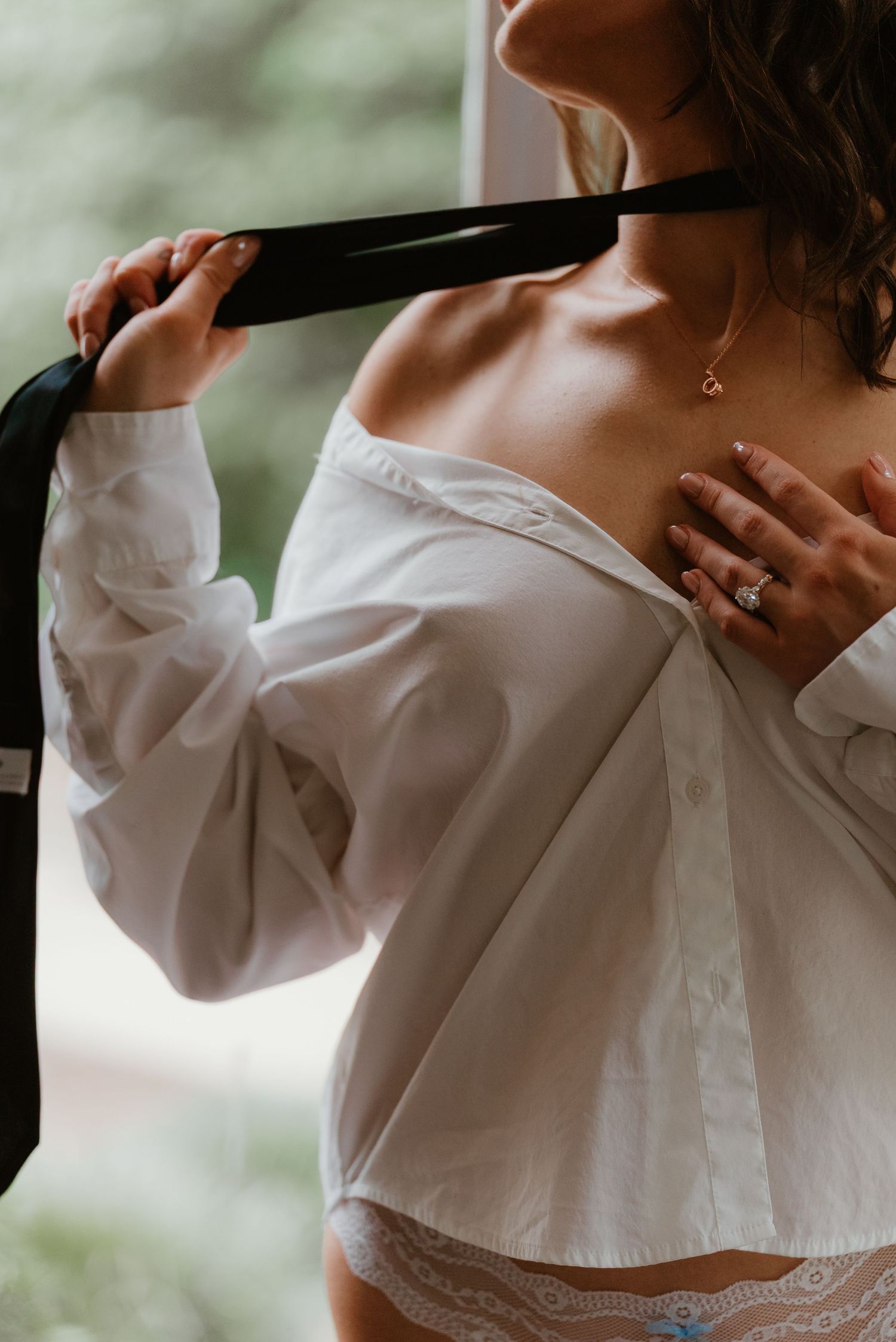 Woman in white off-shoulder shirt holding black tie with diamond engagement ring.