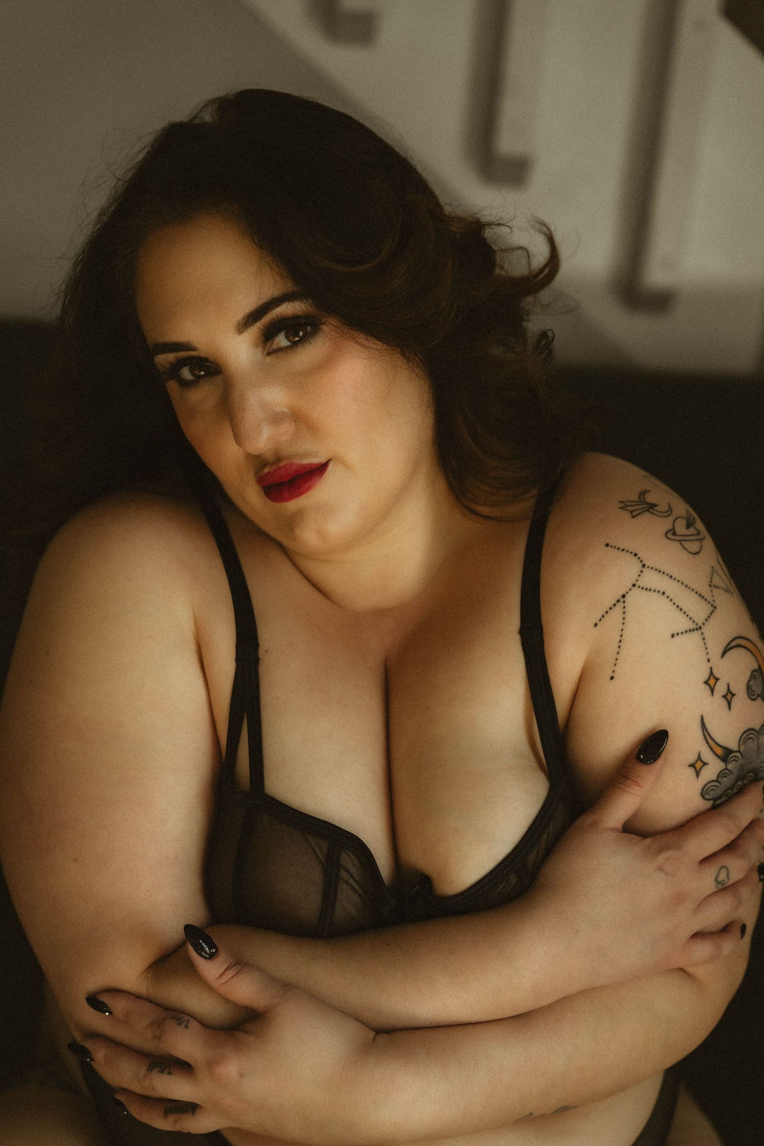 Plus-size woman in sheer black lingerie with constellation tattoo posing with crossed arms.