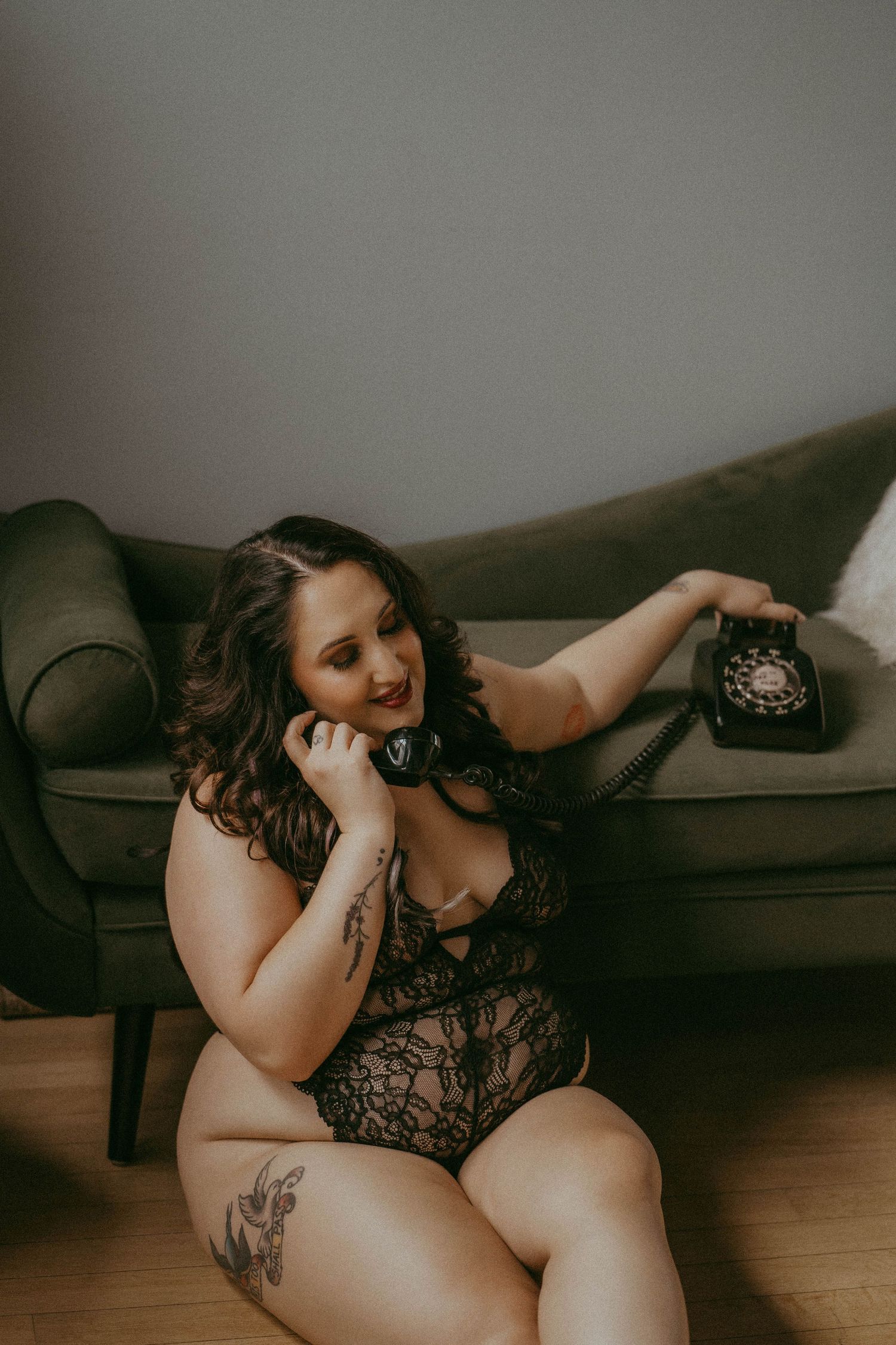 A plus-size woman in black lace bodysuit sits on the floor holding a vintage rotary telephone to her ear.