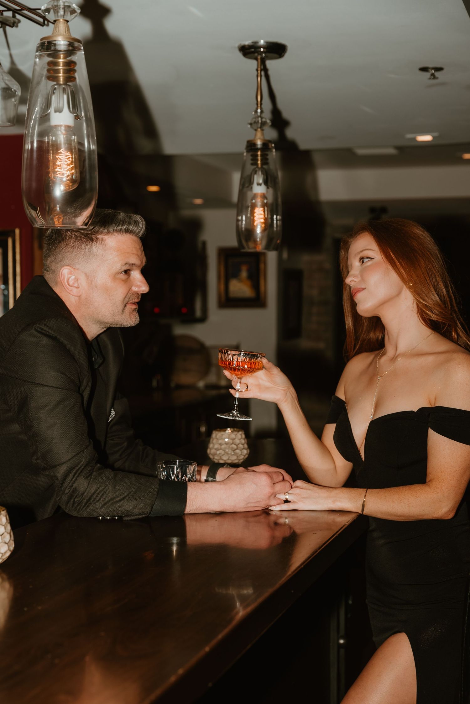 Couple in elegant black attire toasting cocktails at a stylish bar with pendant lighting.