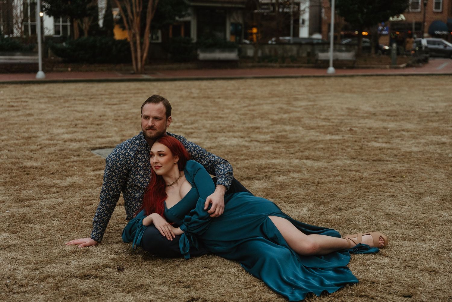 Couple relaxing on dry grass, woman in teal dress leaning against man in floral shirt outdoors.