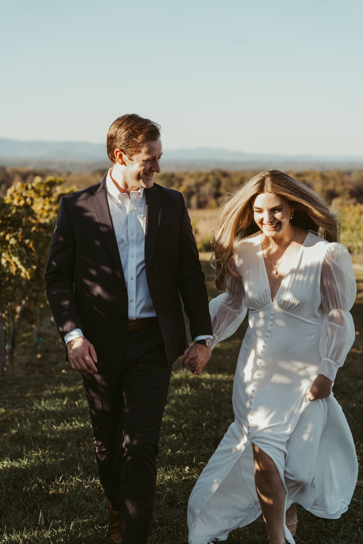 Happy couple walking hand in hand through a vineyard in wedding attire at sunset.