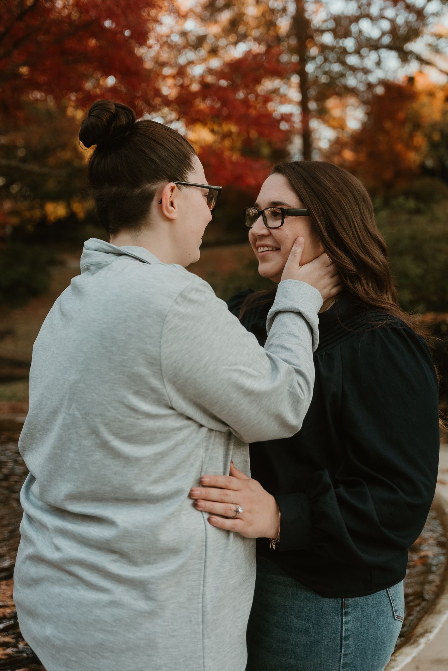 Two women smiling and embracing outdoors surrounded by vibrant red autumn foliage.
