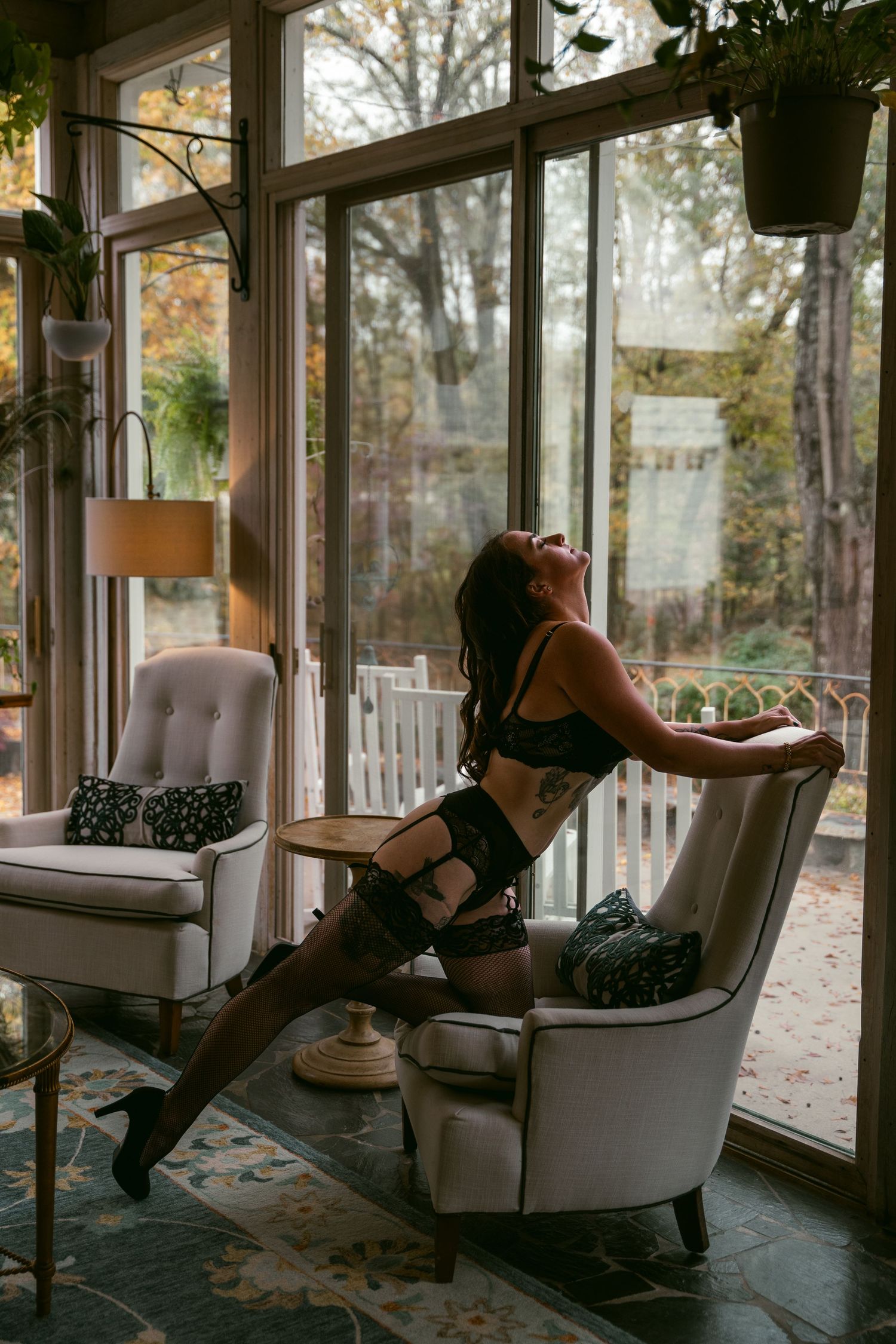 Woman in black lingerie and stockings posing on chair in sunlit room with plants.