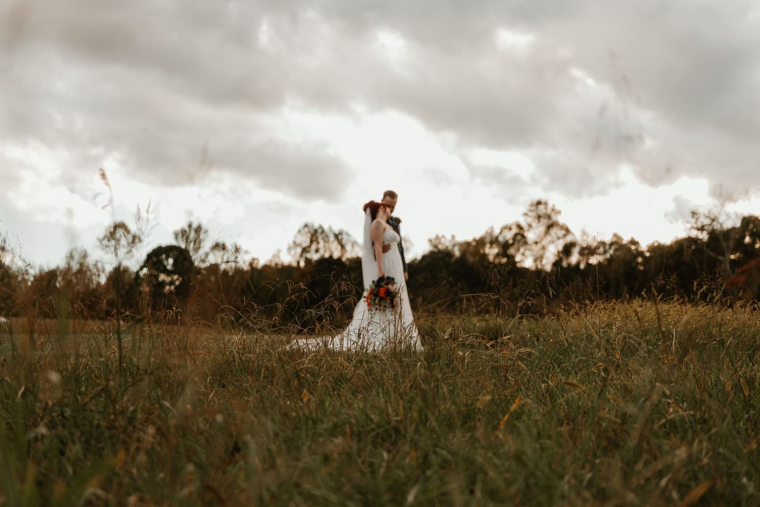Bride and groom stand together in lush green field under moody cloudy sky with bouquet.