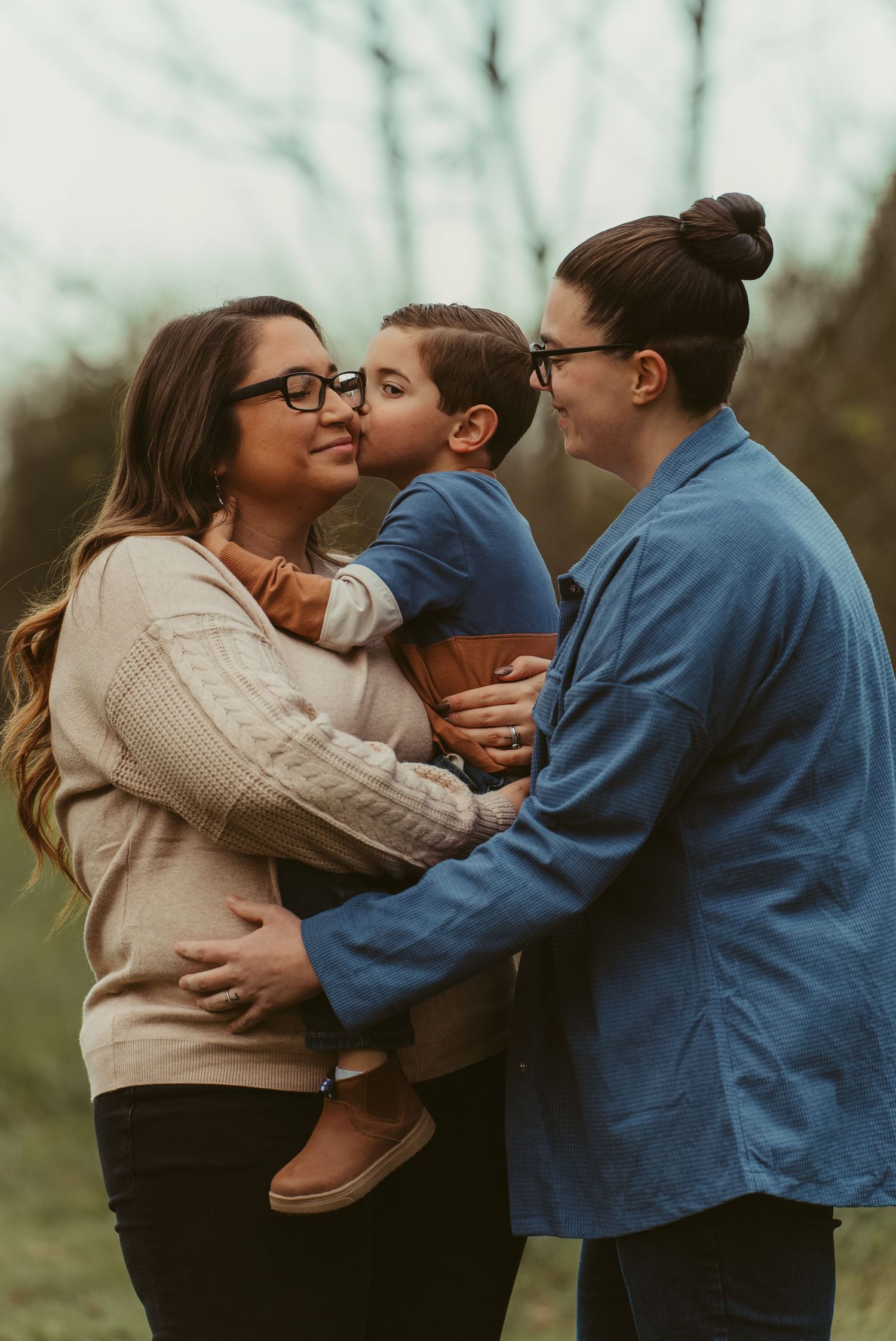 Two mothers share a joyful moment as their young son kisses one mom on the cheek outdoors.