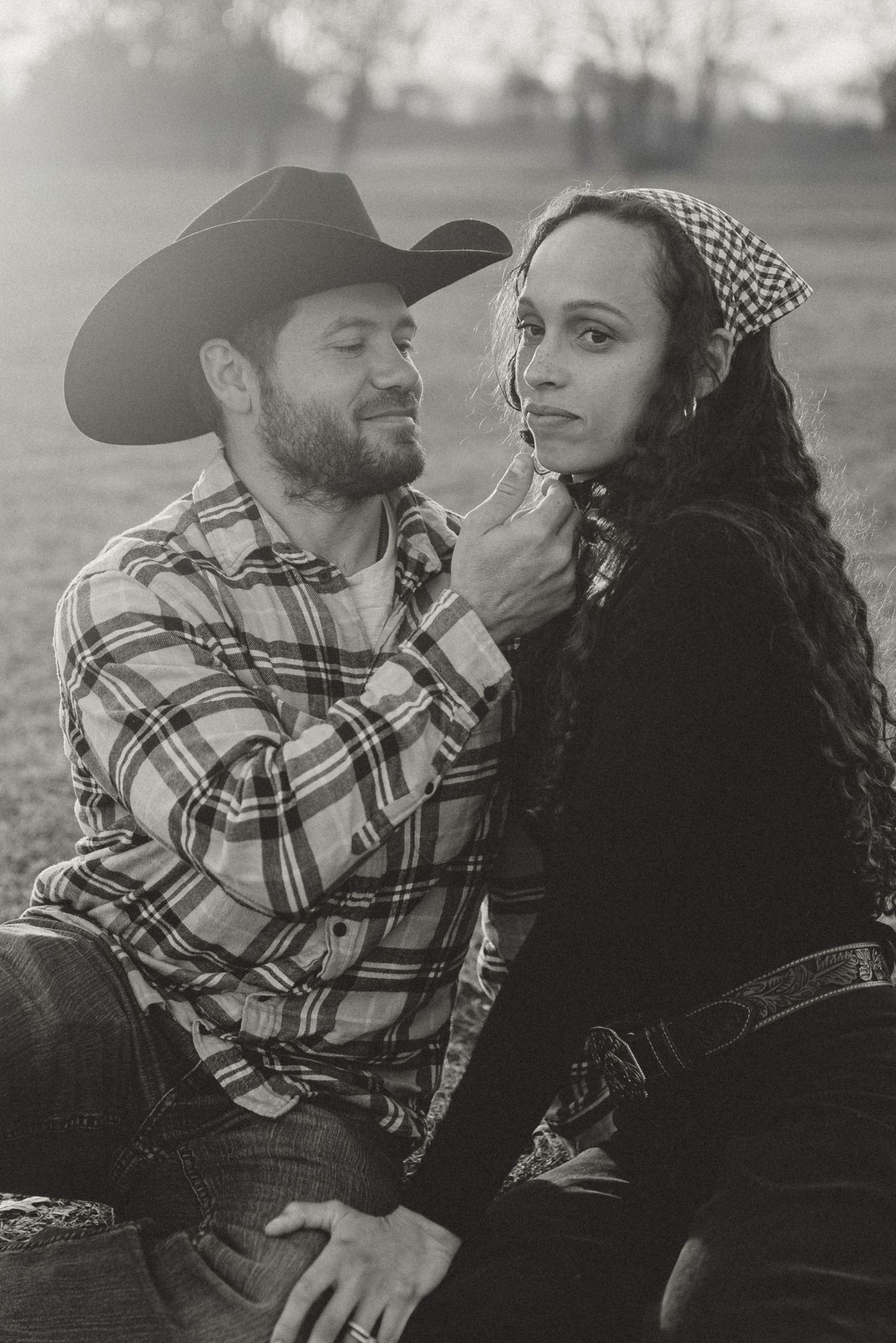 Black and white portrait of a couple sitting together in a field, man in cowboy hat and plaid shirt.