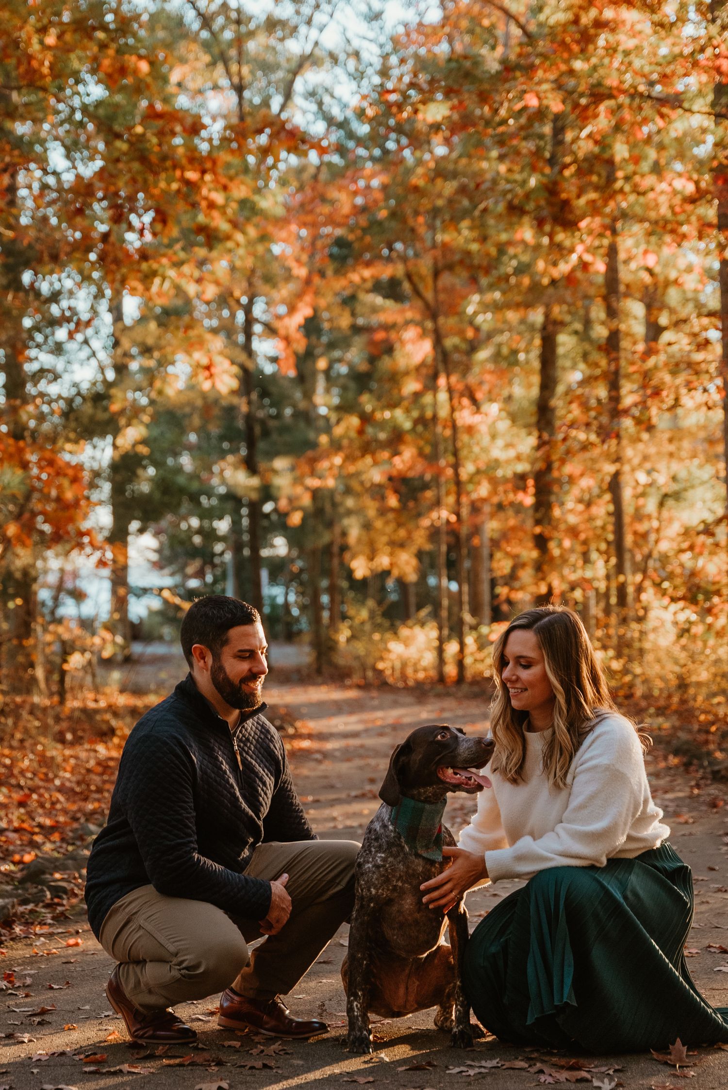 Smiling couple crouching with their German Shorthaired Pointer dog on a colorful autumn forest path.