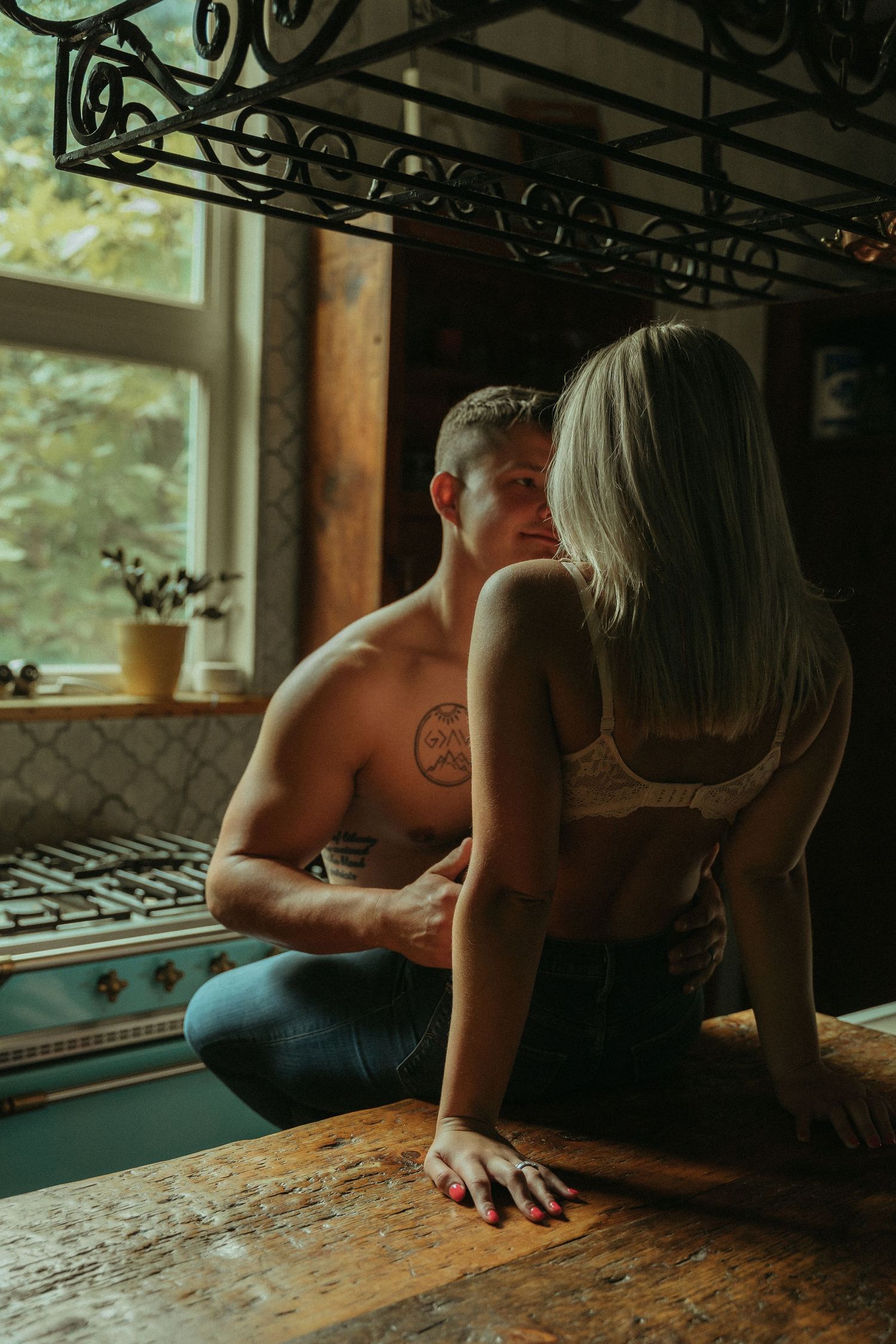 Shirtless tattooed man and blonde woman in lace bralette share intimate moment by kitchen window.