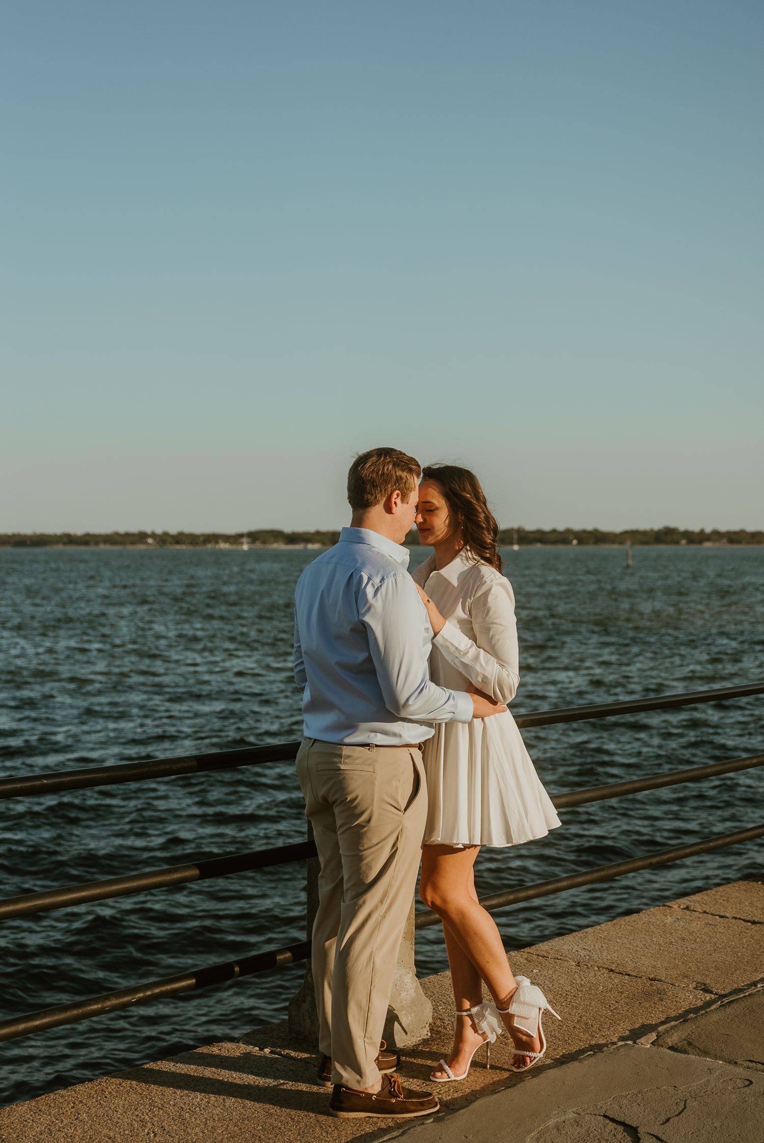 Couple nearly kissing while standing on a waterfront pier during golden sunset.