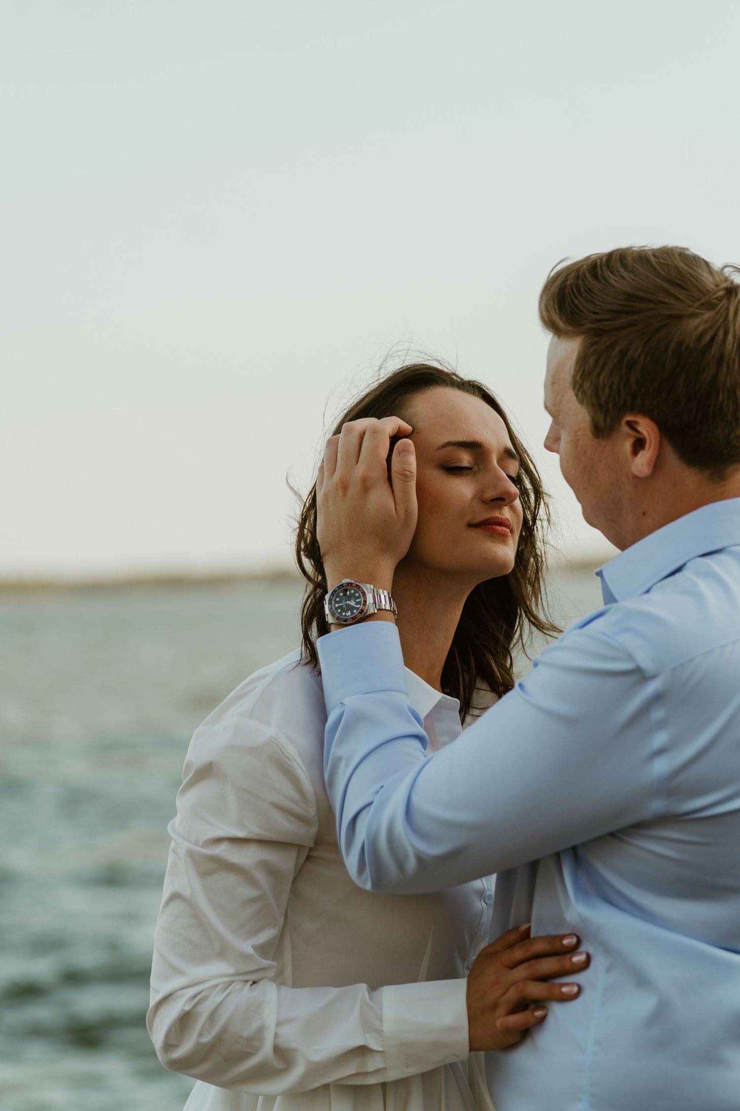 Man tenderly brushing hair from woman's face near the water during a romantic sunset couples photo session.