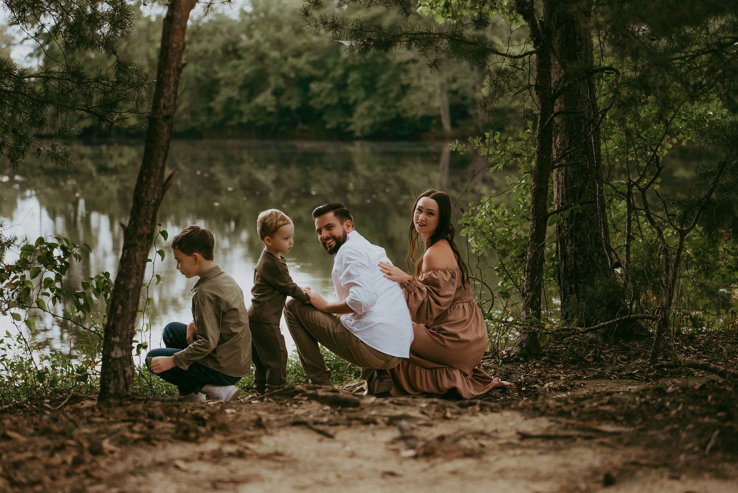A family of four sits together on a wooded riverbank surrounded by lush green trees.