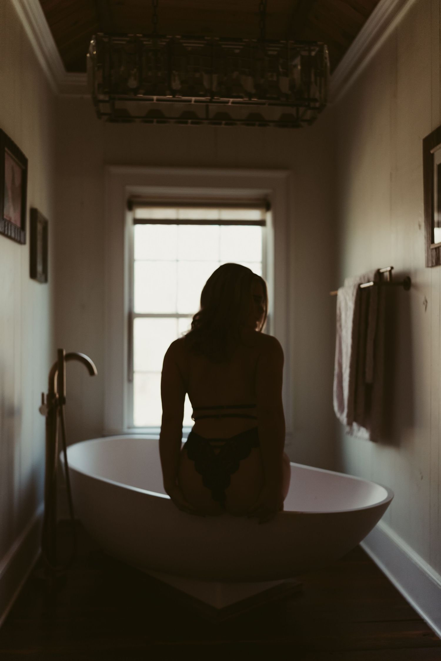 Silhouette of a woman sitting on a freestanding bathtub facing a window in a dimly lit bathroom.