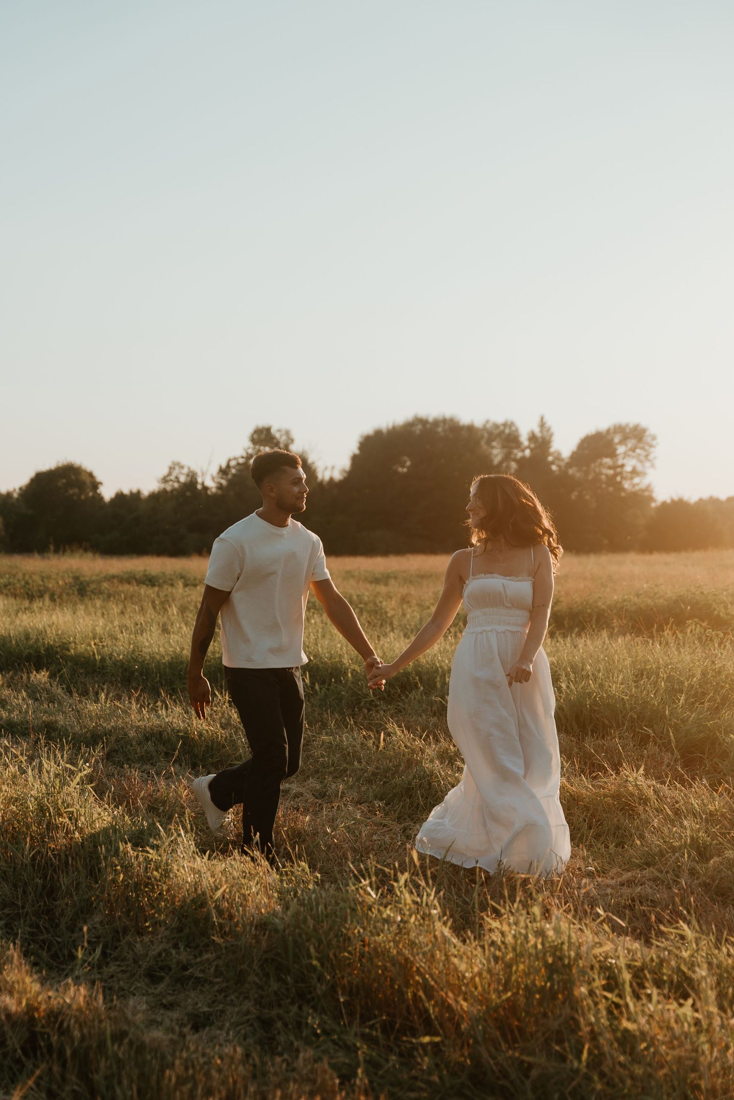 Engaged couple walking hand in hand through a sunlit meadow during beautiful golden hour.