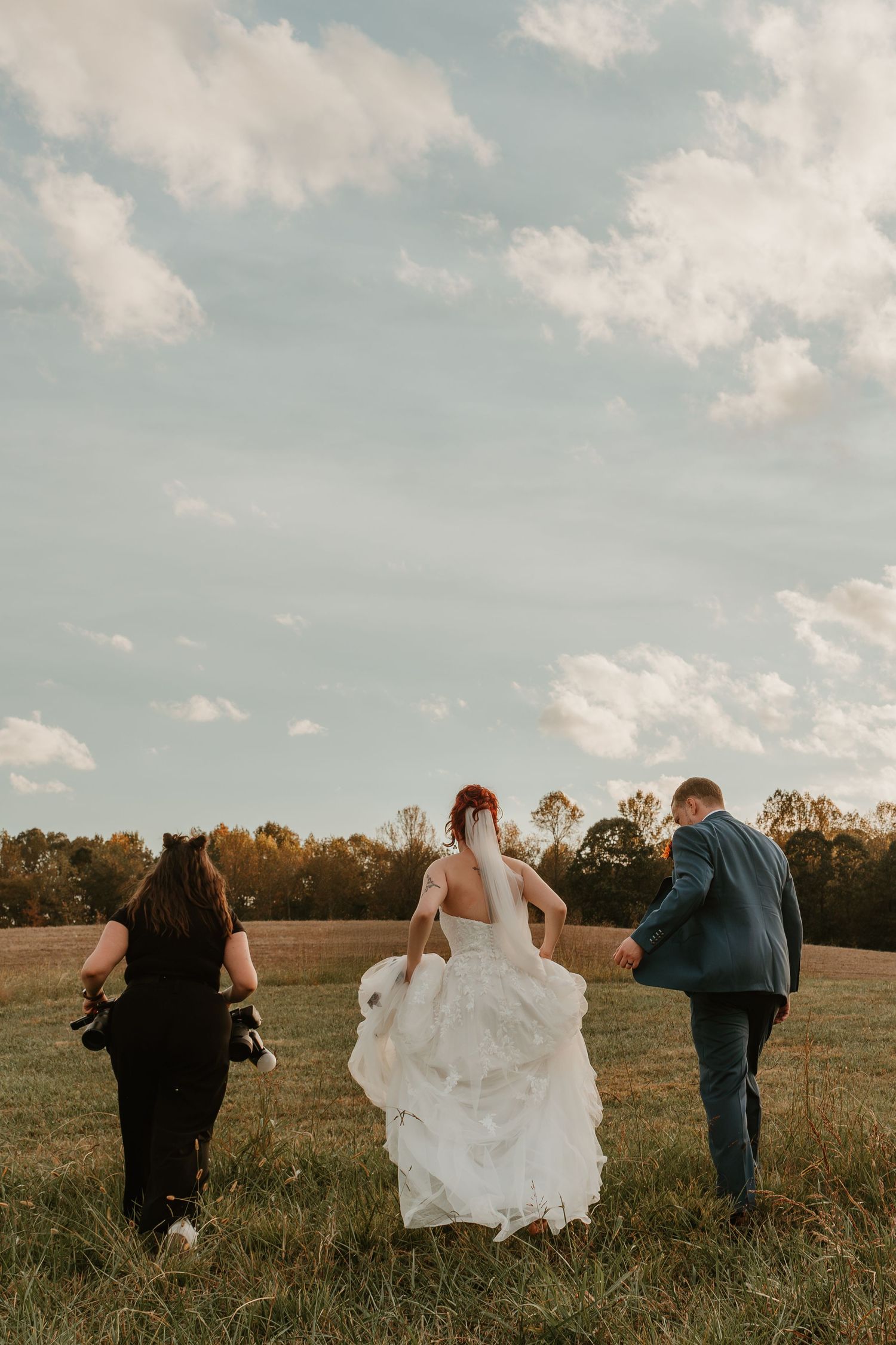 Wedding photographer walking with bride and groom across open field under dramatic cloudy sky.