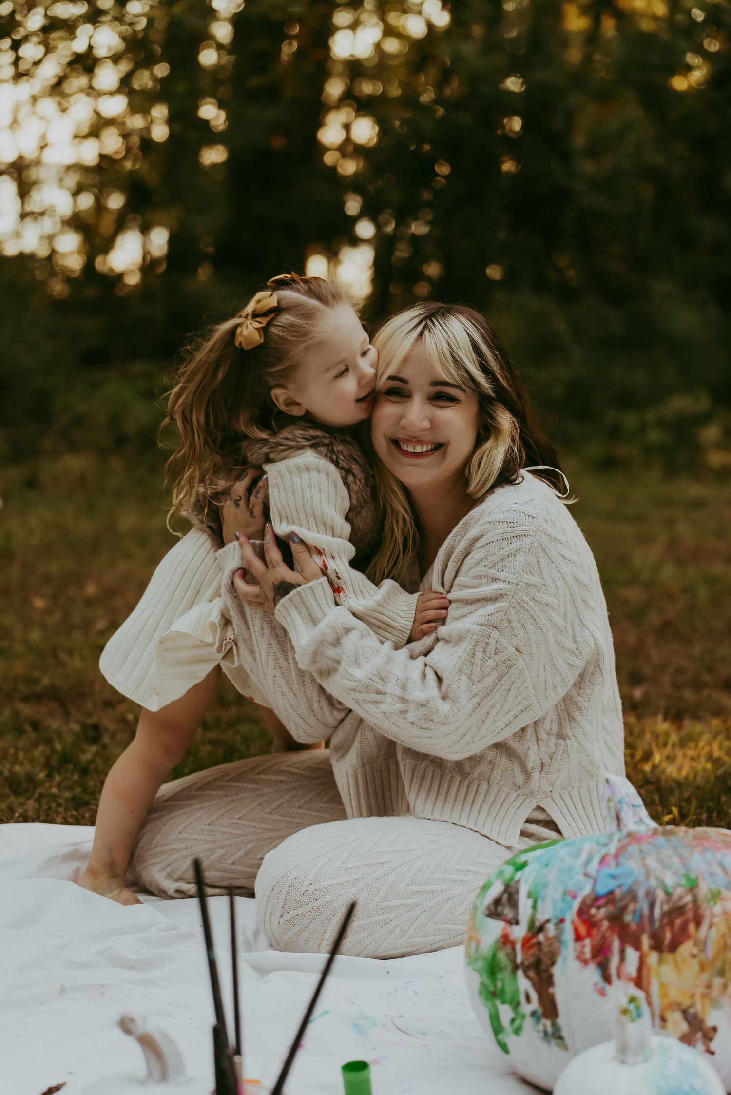A smiling woman in a cream sweater sits on a blanket outdoors while a young girl kisses her cheek.
