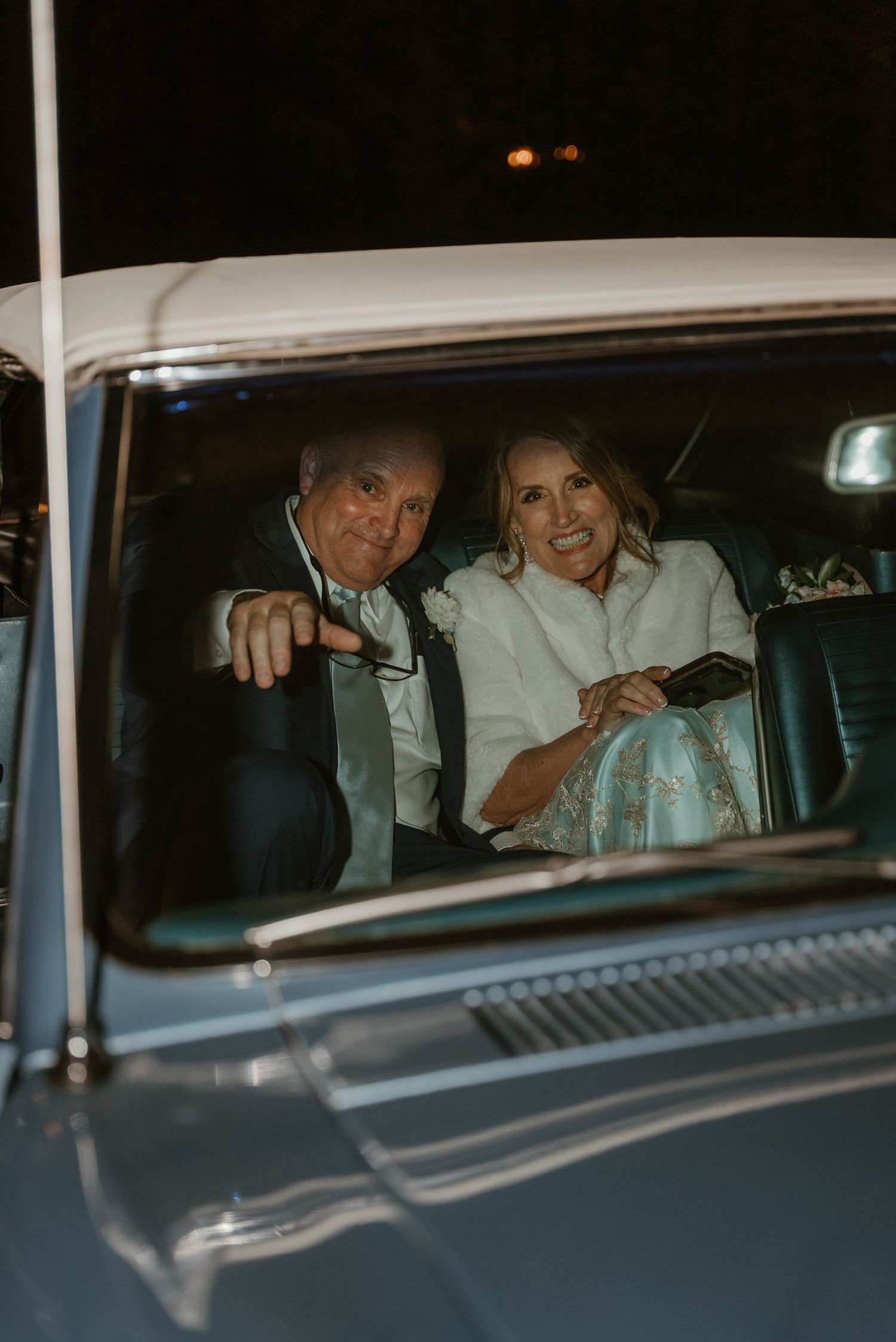 Newlywed couple smiling inside a classic vintage car at night, bride wearing a white jacket.