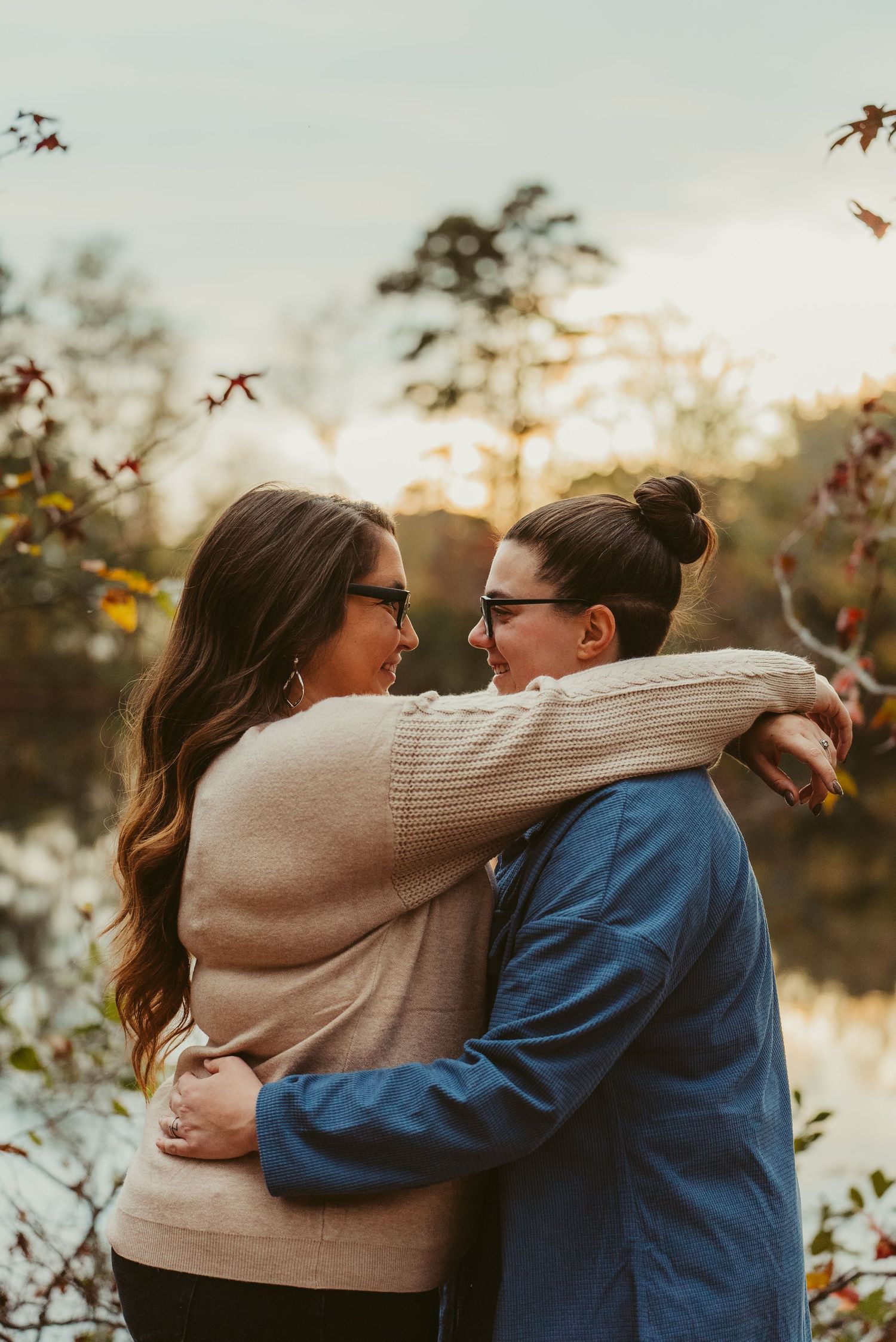 Two women embracing and smiling at each other near a lake at sunset.