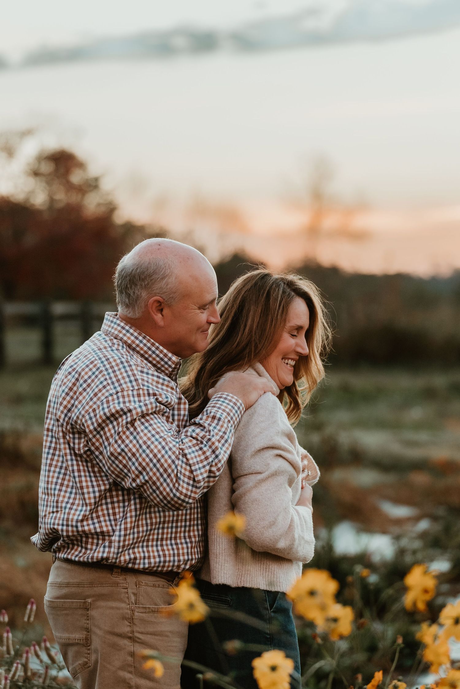 Couple embracing in a golden field of yellow flowers at sunset during fall engagement session.