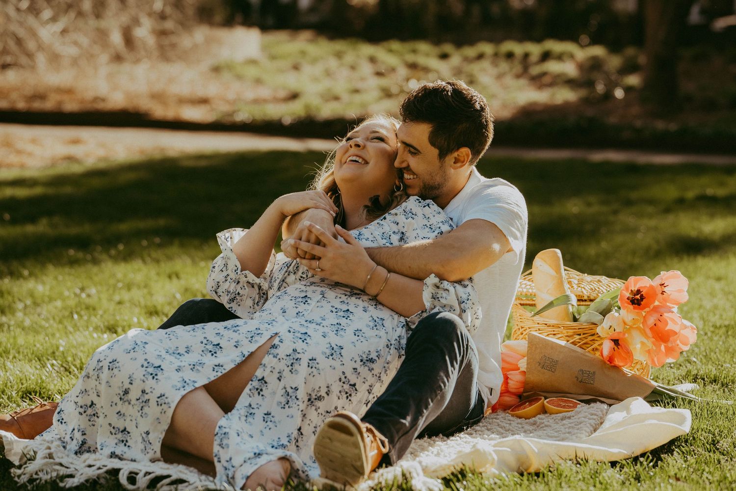Happy couple laughing together on a picnic blanket in a sunny park with flowers nearby.