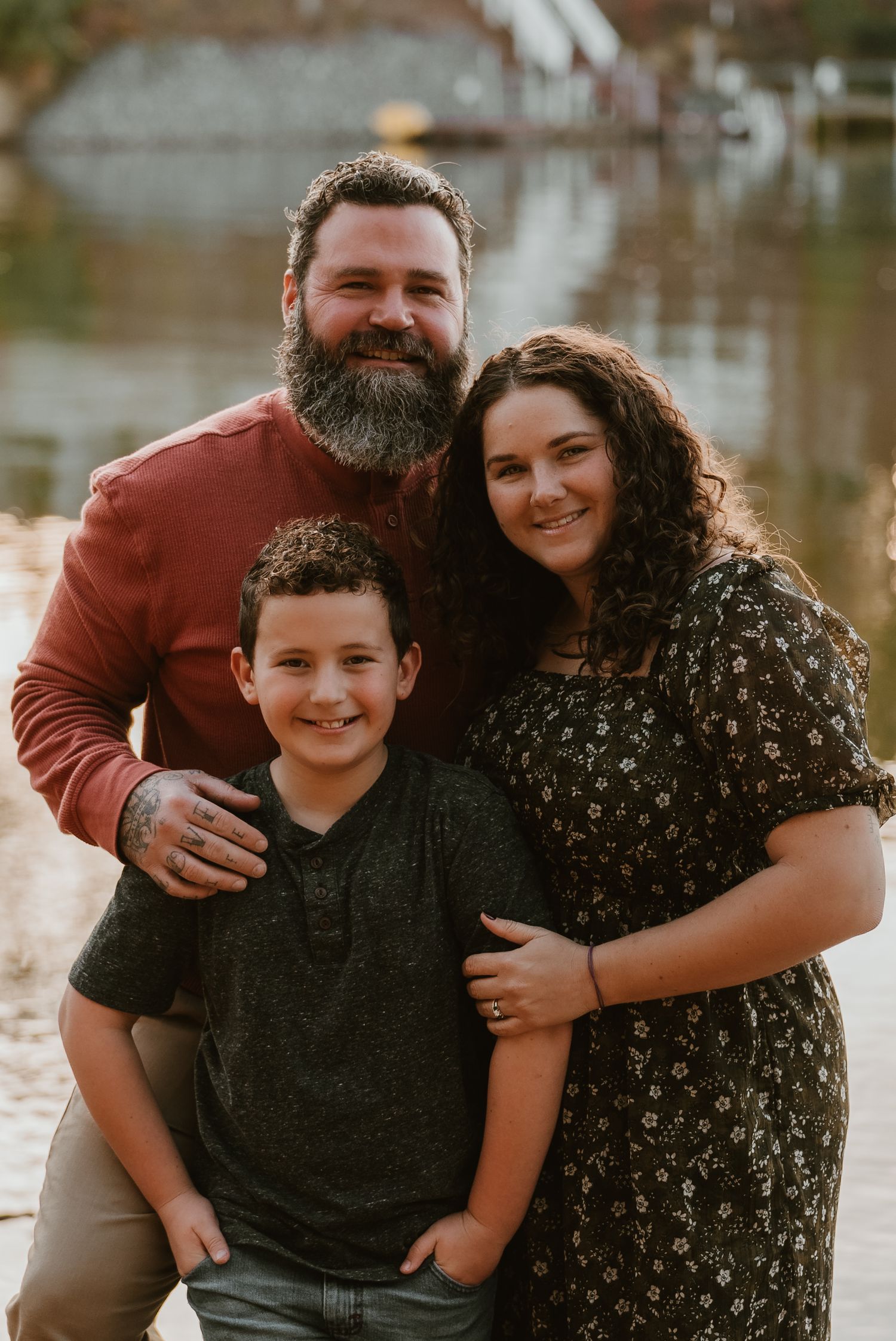 A bearded father, curly-haired mother, and young son smile together in a warm outdoor portrait.