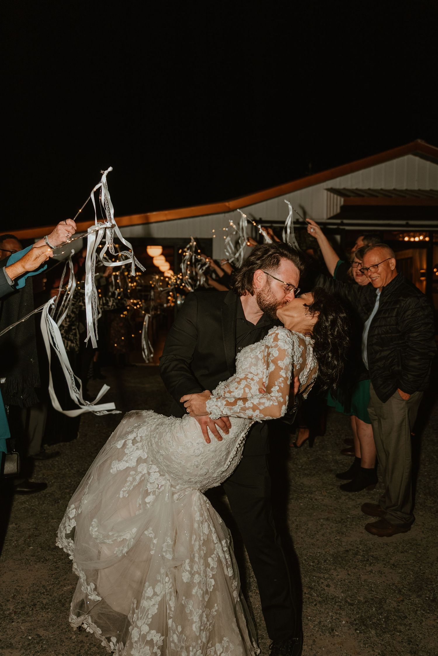 Bride and groom sharing passionate dip kiss during outdoor wedding send-off with ribbon wands at night.
