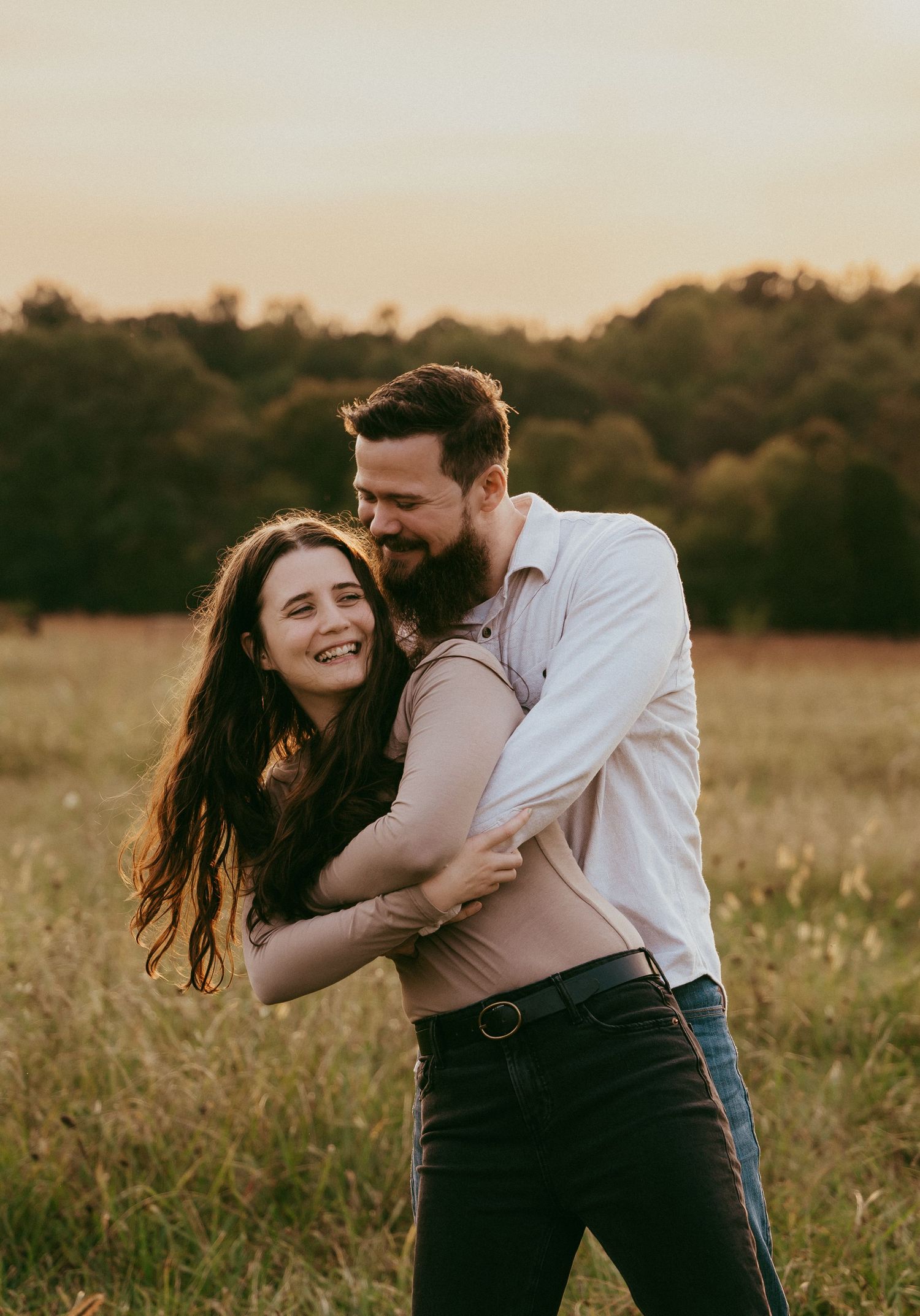 Couple embracing and laughing in a golden sunset field with trees in background.