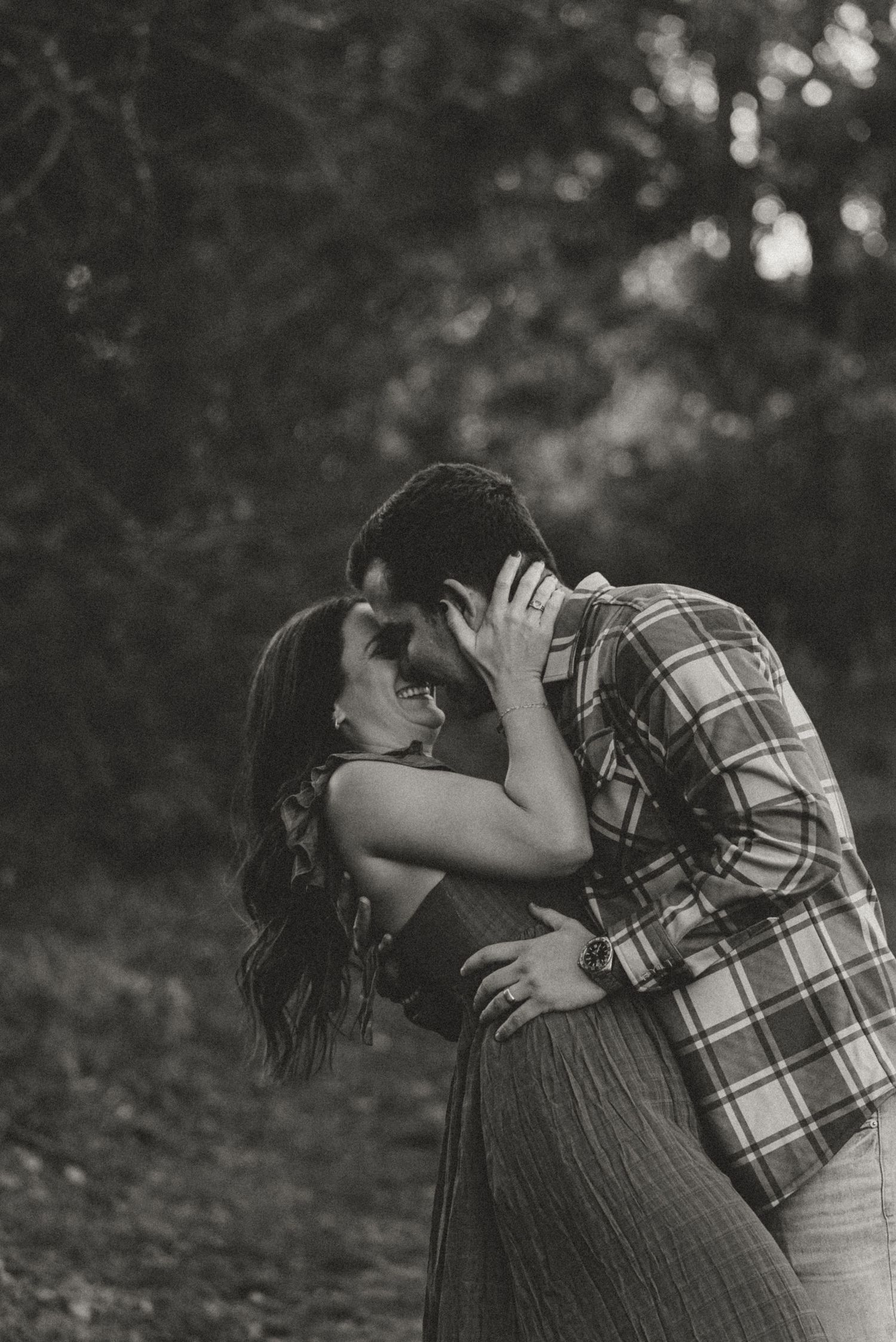 Black and white photo of a couple embracing and laughing together in a wooded outdoor setting.