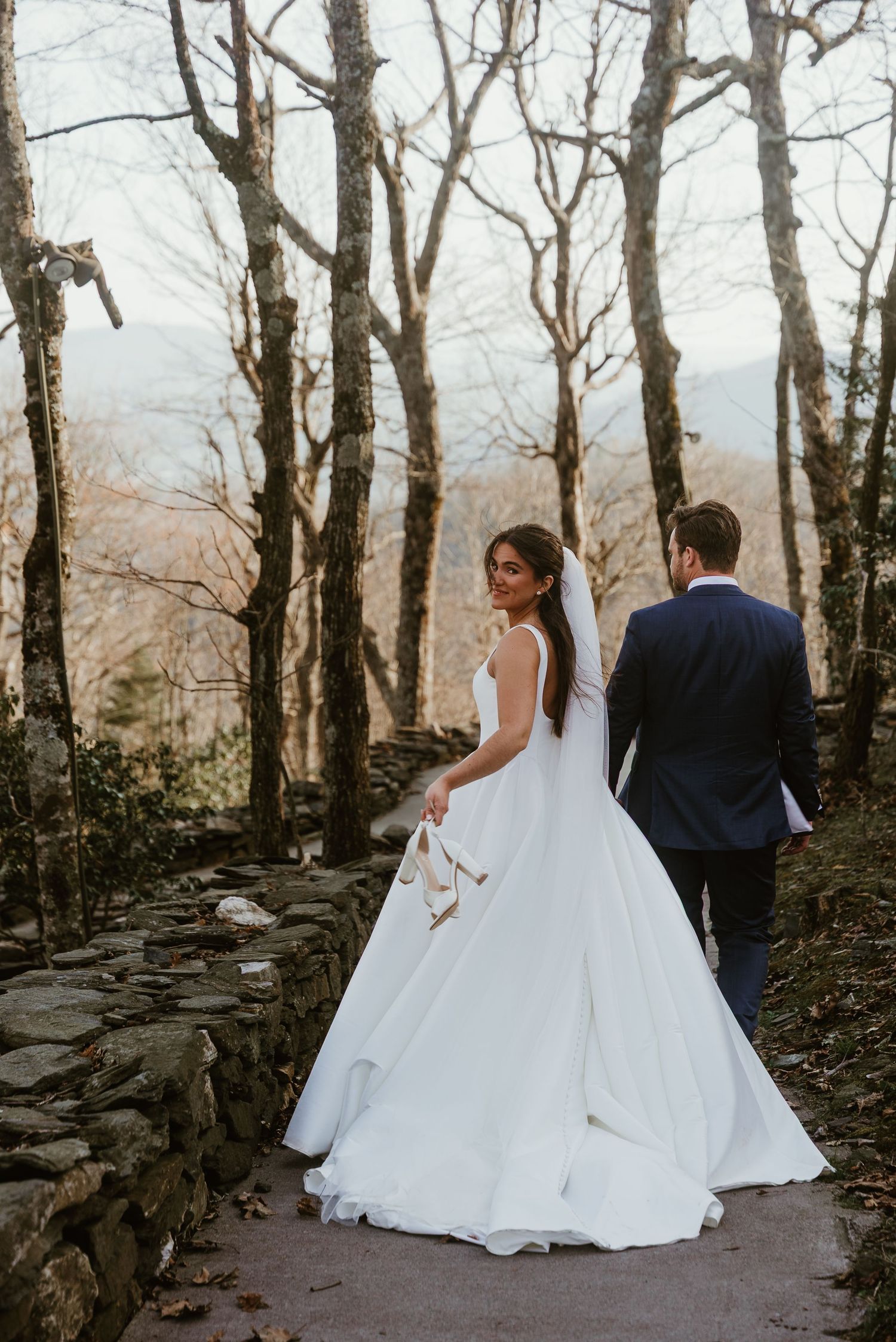 Bride in ballgown looks back while walking with groom through a wooded stone path with mountain views.