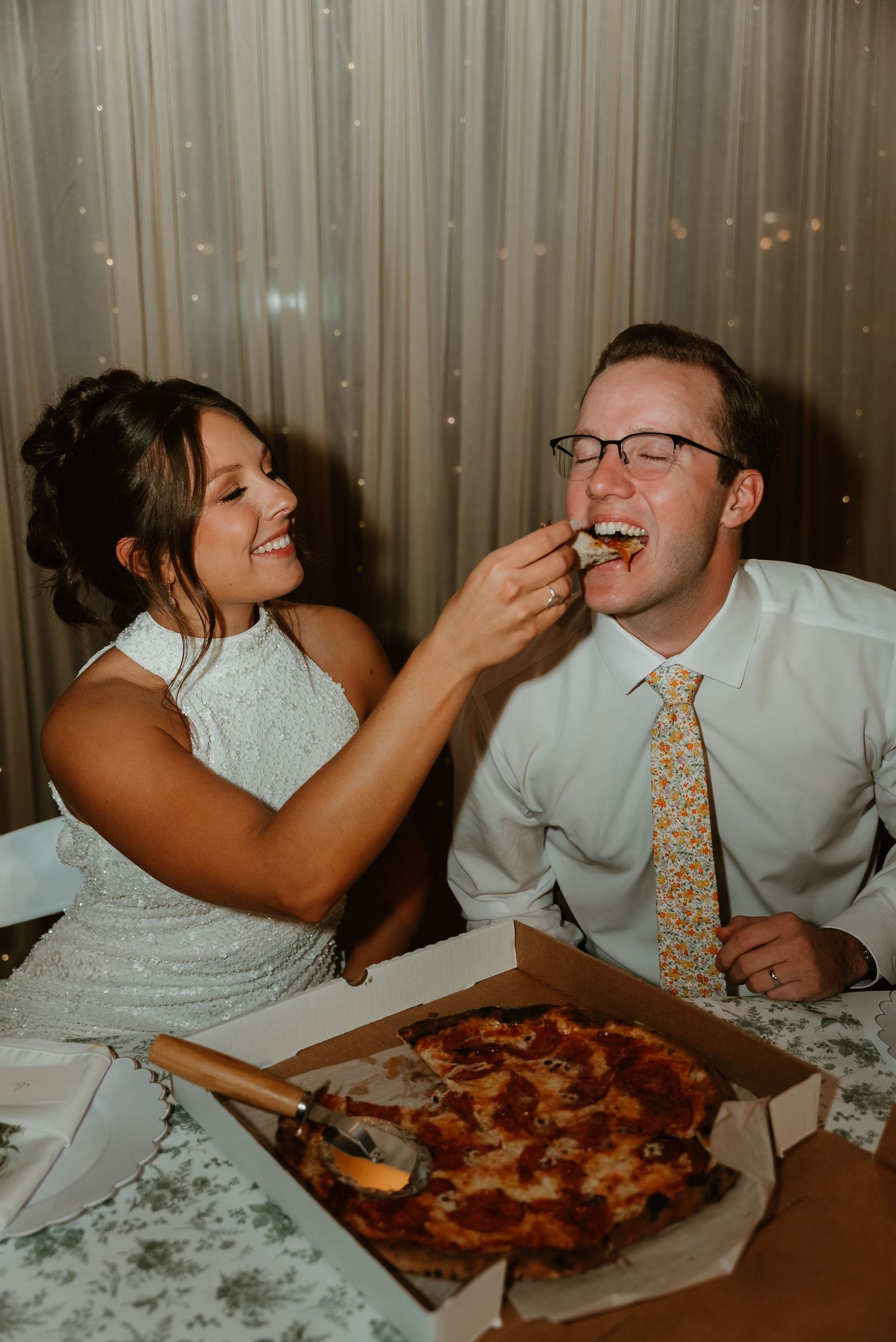Smiling bride feeding groom pizza at wedding reception table with floral tablecloth and bokeh lights.