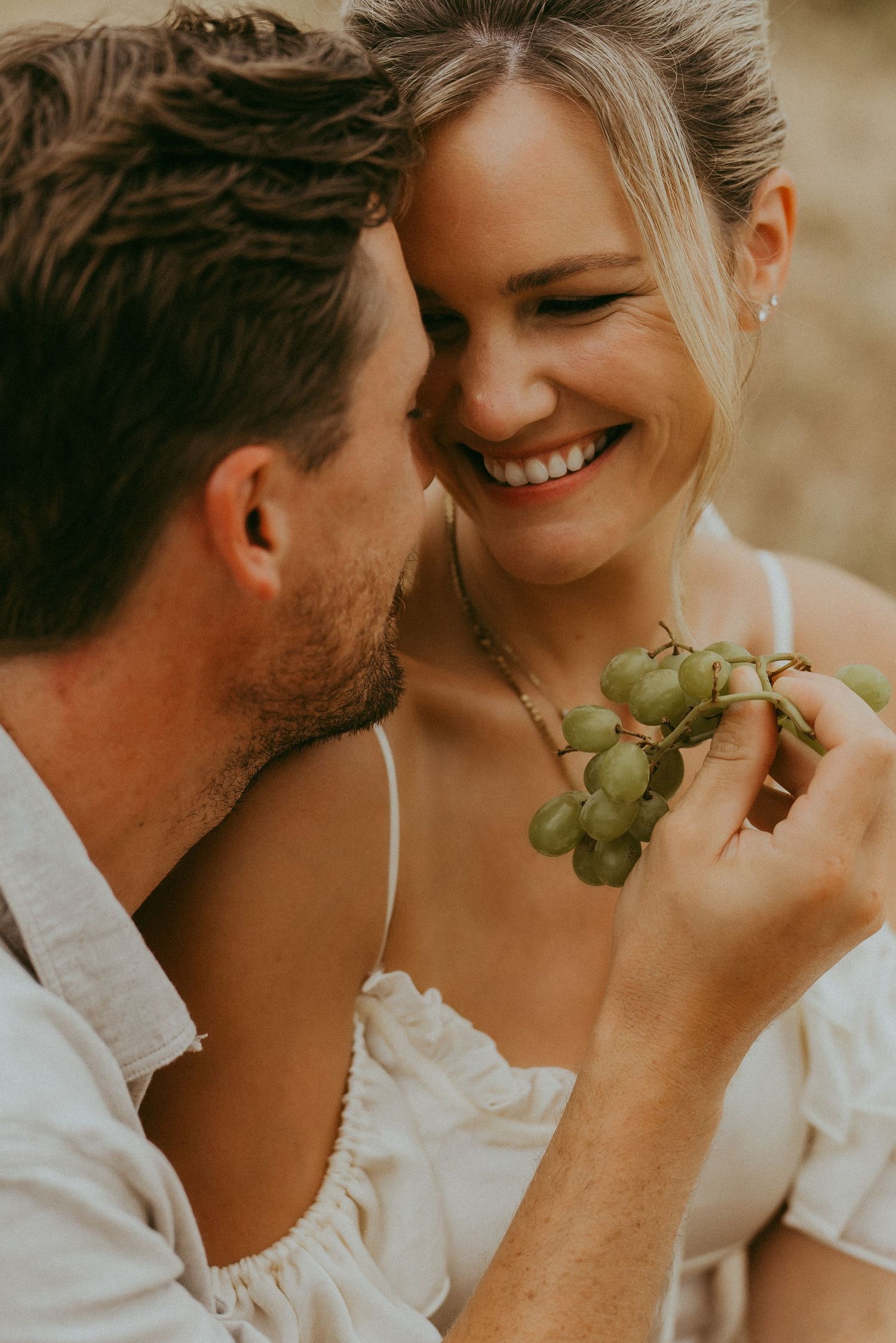 Smiling couple sharing green grapes during a romantic outdoor picnic engagement photo session.