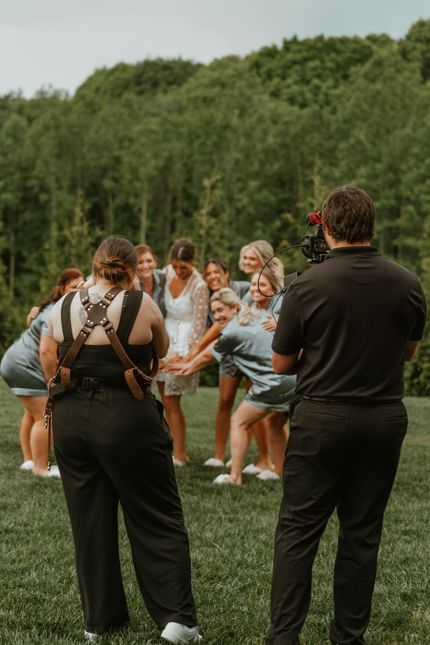 Wedding photographer and videographer capturing bridesmaids group photo on green lawn with forest backdrop.