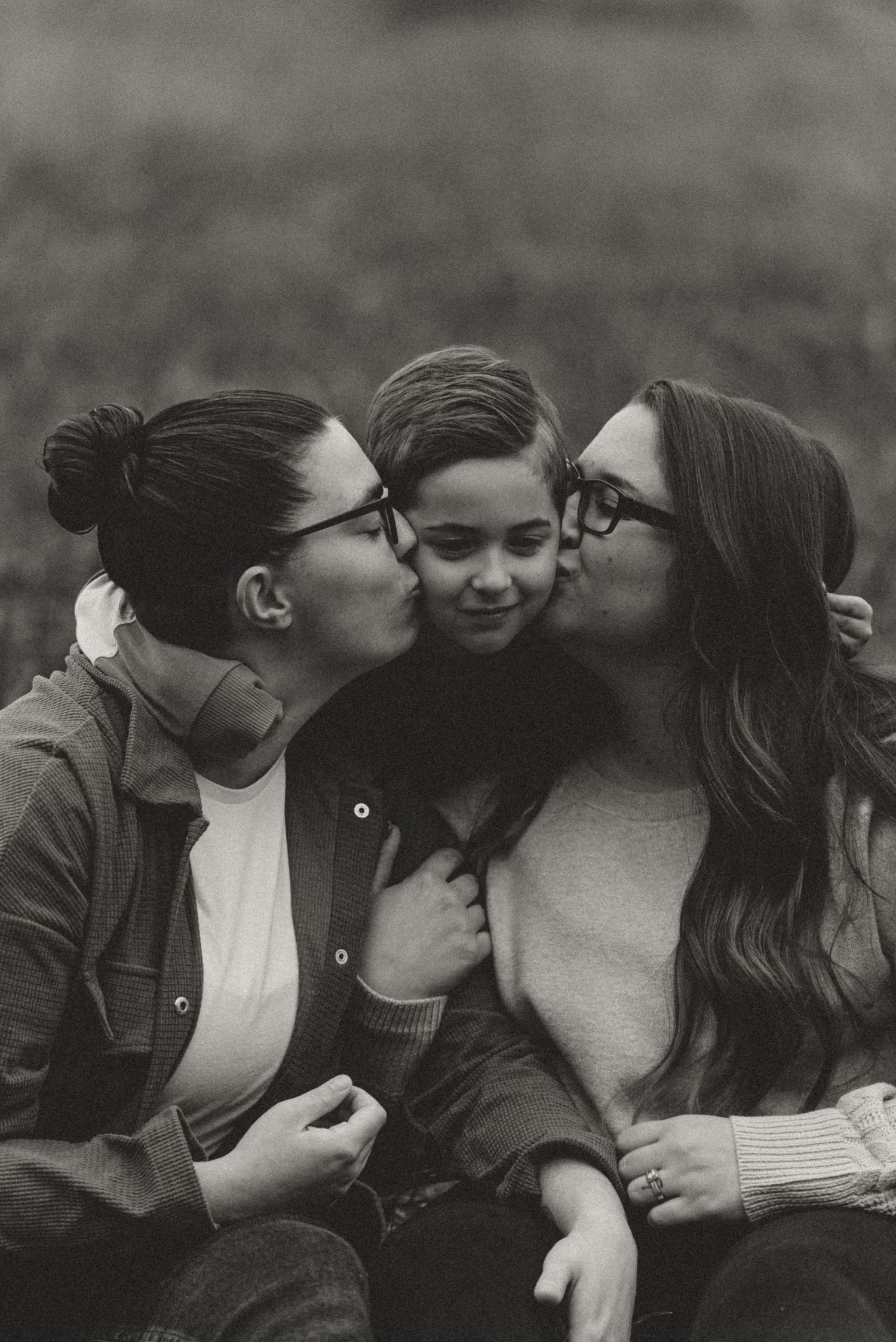 Black and white photo of two mothers kissing their young son on the cheeks outdoors.