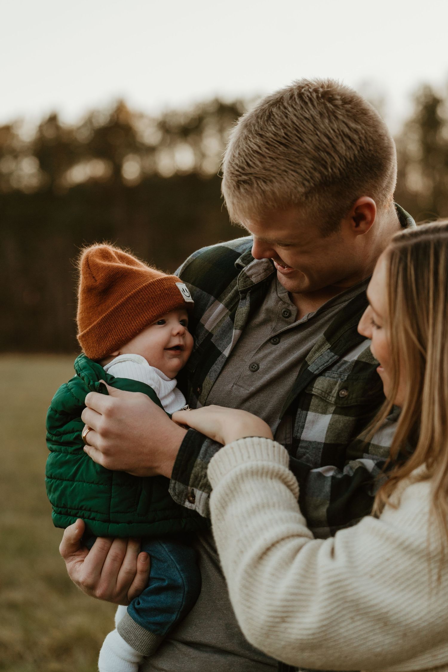 Young parents smiling and cradling their newborn baby wearing an orange knit hat outdoors.
