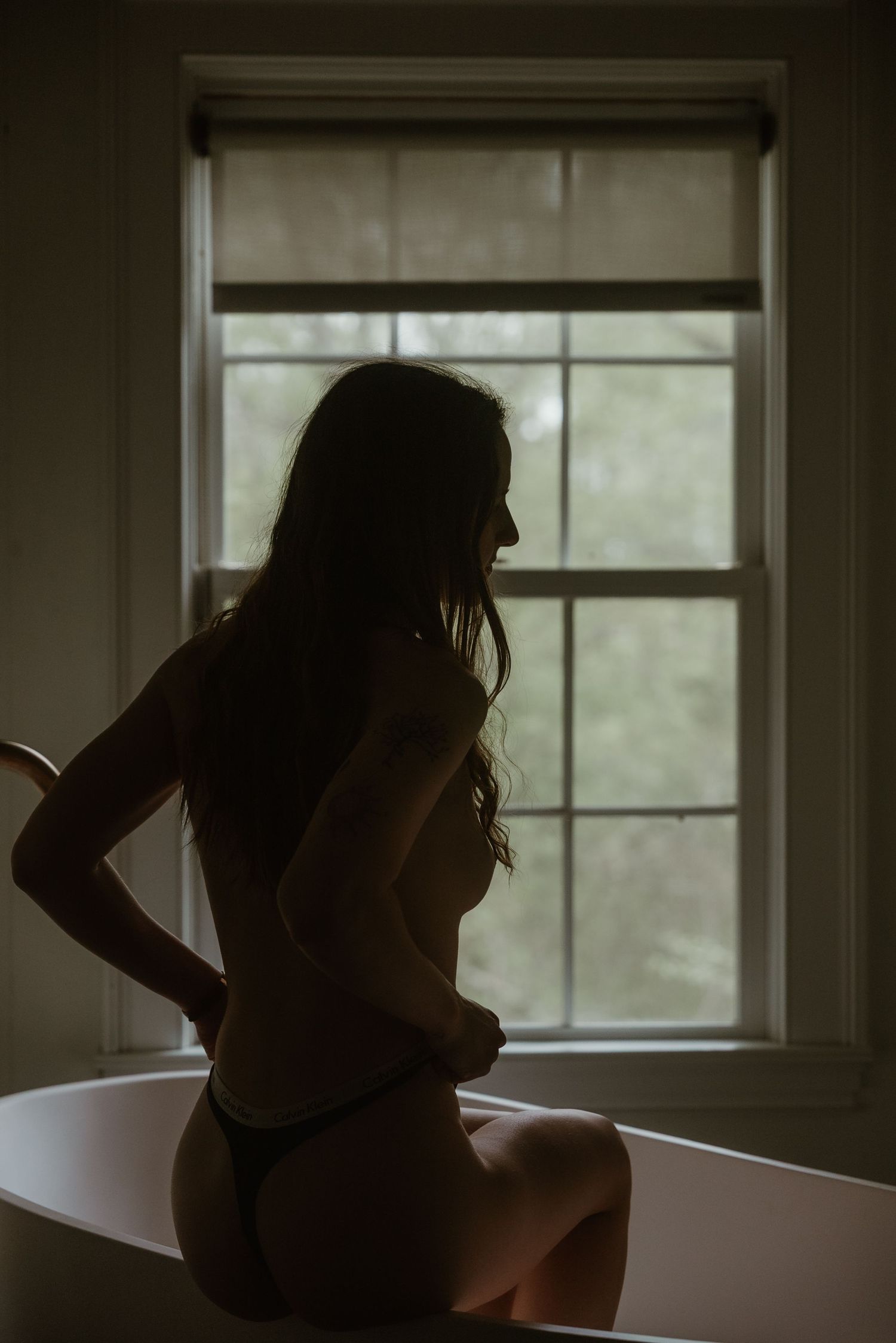 Silhouette of a woman in lingerie perched on a bathtub edge by a window.