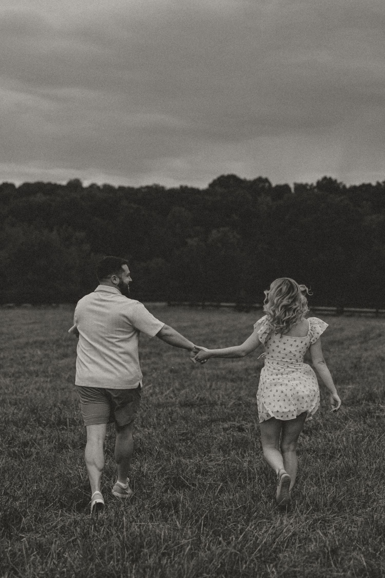 Black and white photo of couple holding hands running through an open grassy field.