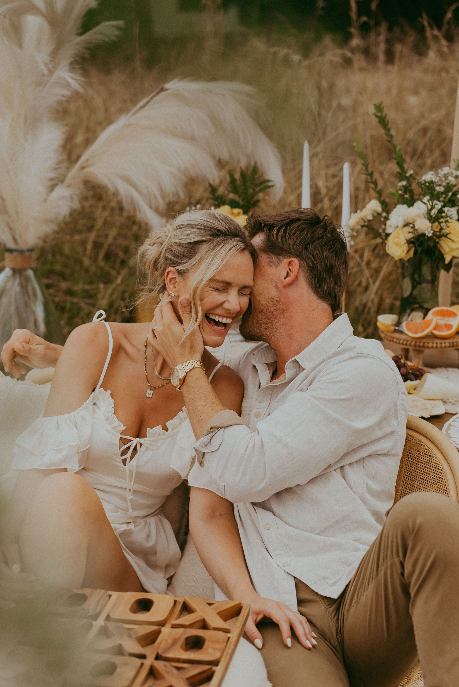 Joyful couple laughing together at a boho outdoor picnic surrounded by pampas grass and flowers.