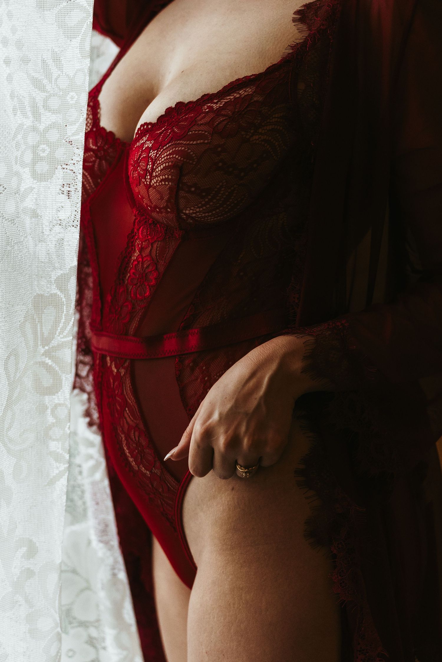 Close-up of woman wearing red lace lingerie bodysuit standing by window in sensual boudoir photography.