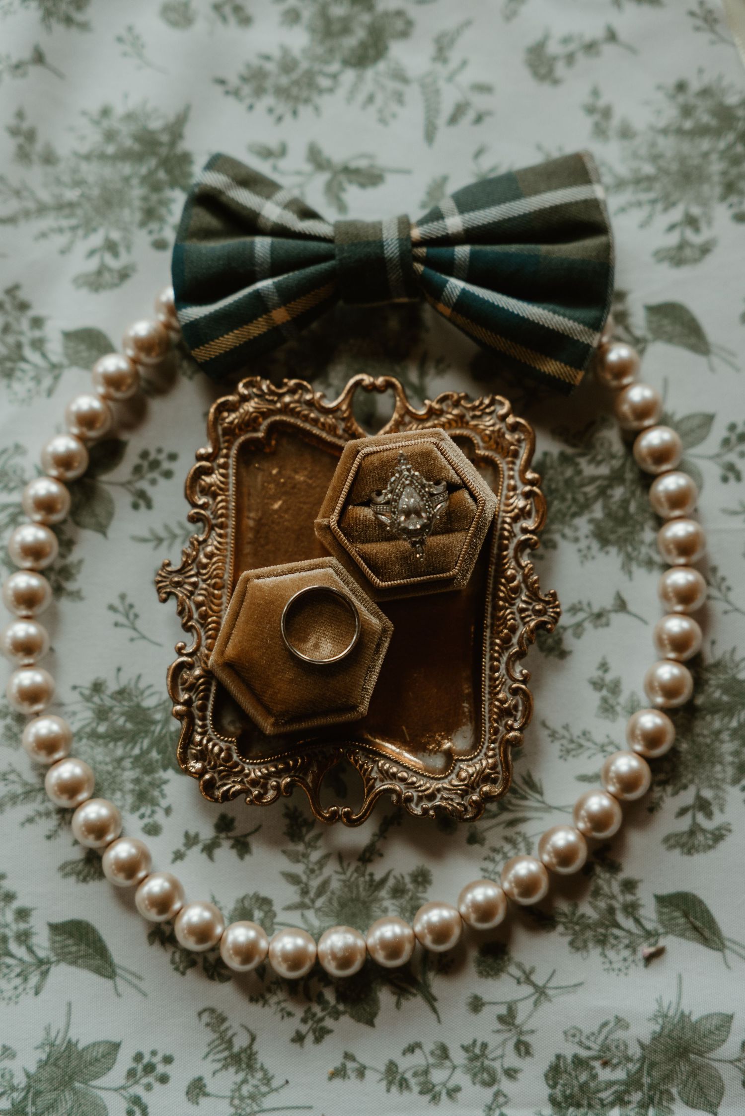 Pearl necklace, wedding rings in gold hexagon box on ornate tray with plaid bow tie on floral background.