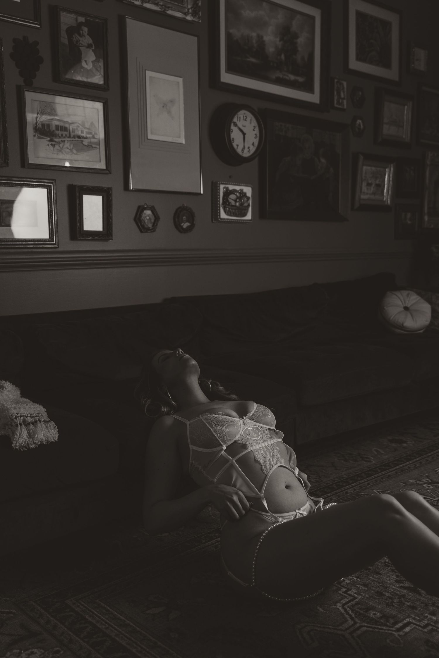 Woman in white lingerie reclines on sofa surrounded by vintage wall art and clock in moody black and white boudoir photo.