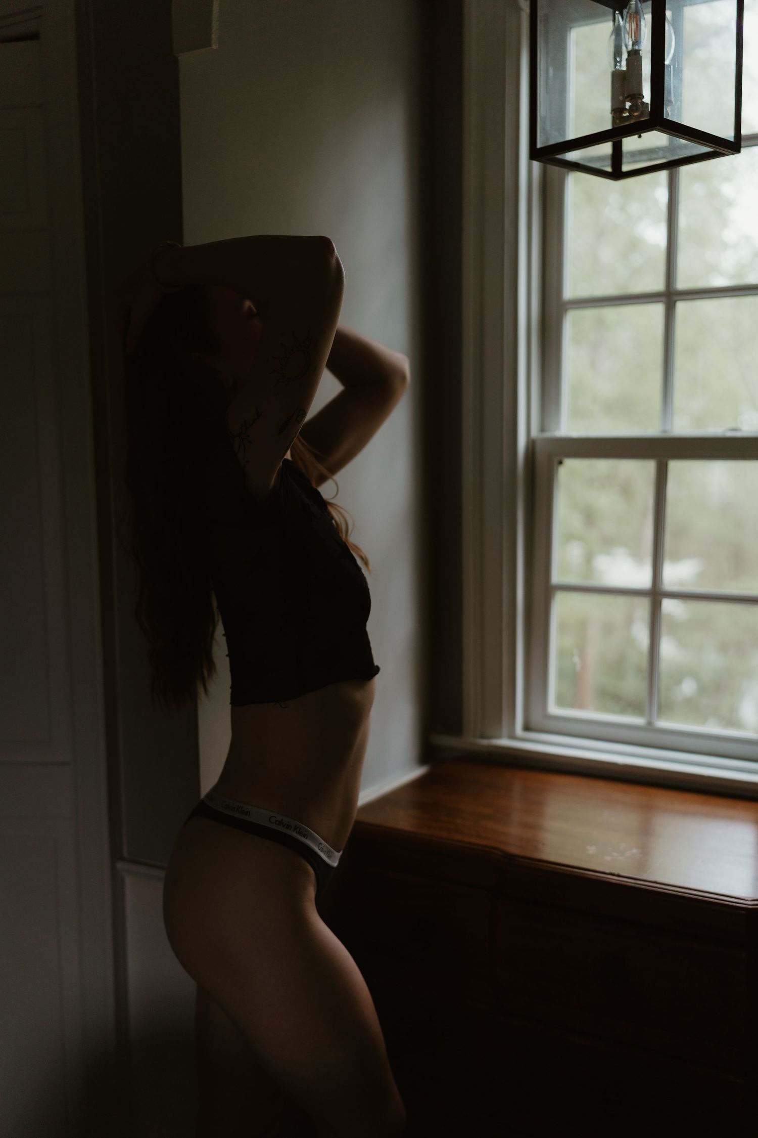 Silhouette of a woman in lingerie standing by a bright window in dim light.