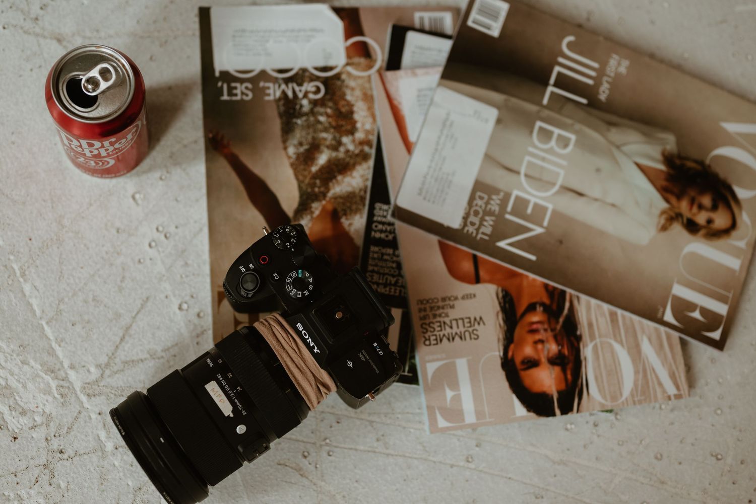 Flatlay of a DSLR camera, Vogue magazines, and a soda can on a marble surface.