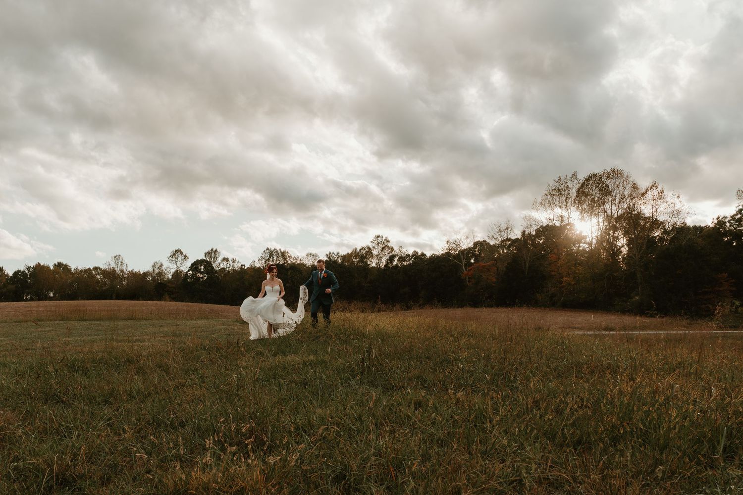 Bride and groom walk hand in hand across open field under dramatic cloudy sky at sunset.