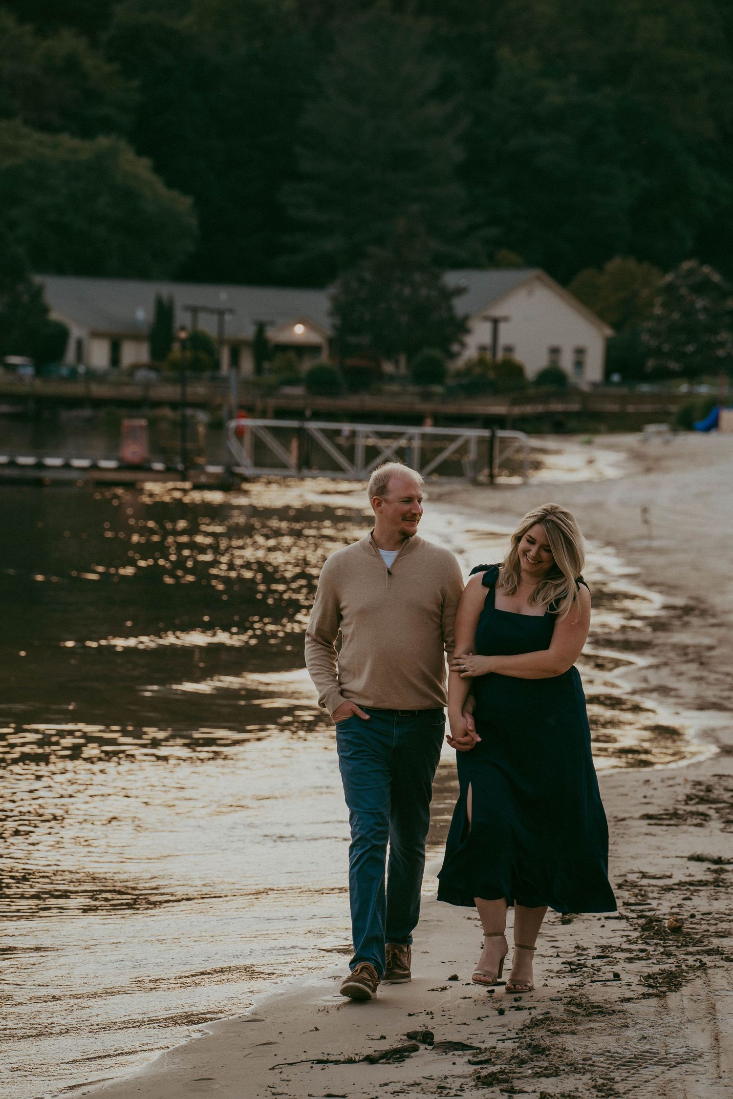 Couple walking hand in hand along a golden sunset shoreline with a marina in background.