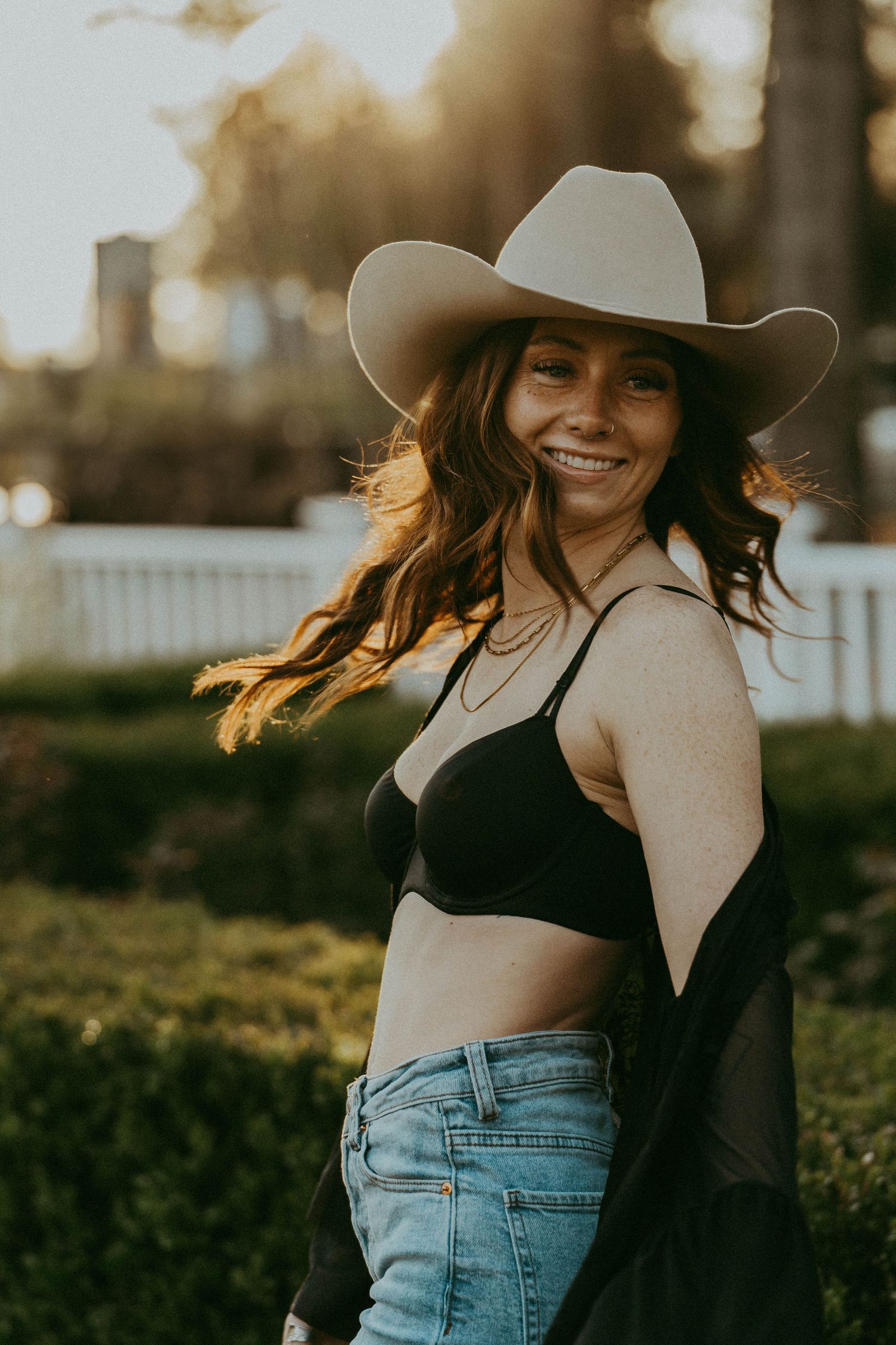 Smiling woman in black bralette and jeans wearing wide-brim cowboy hat in golden sunset light.