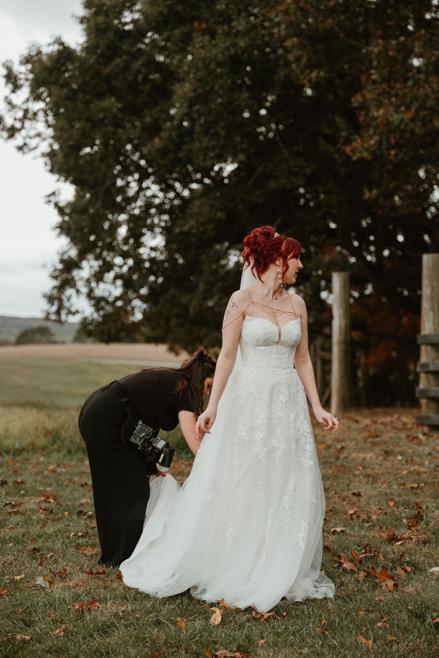 Bride with red hair in white lace wedding gown being helped by assistant outdoors near trees.