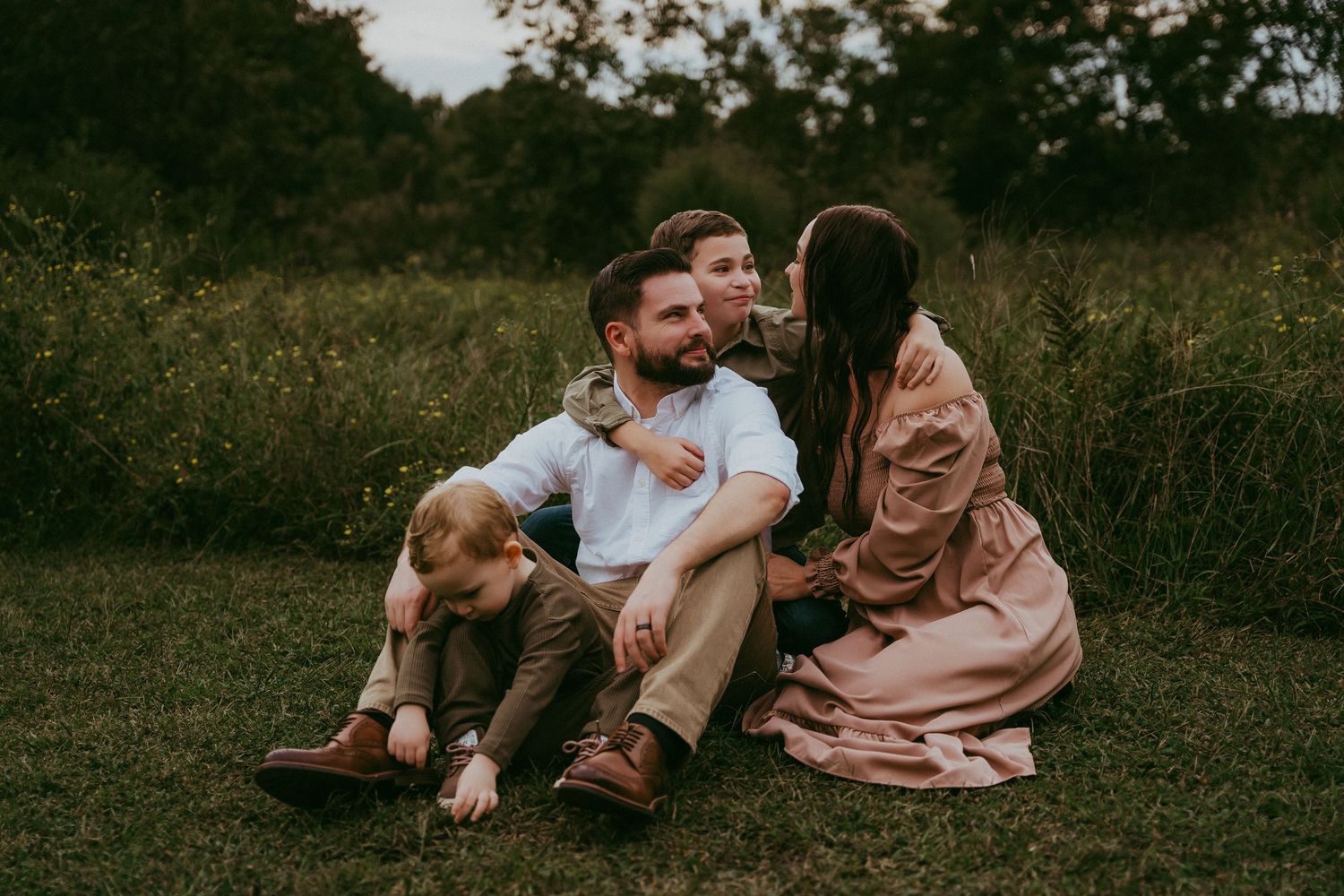 A bearded man sits on grass with two children as a woman in a pink dress leans in affectionately.