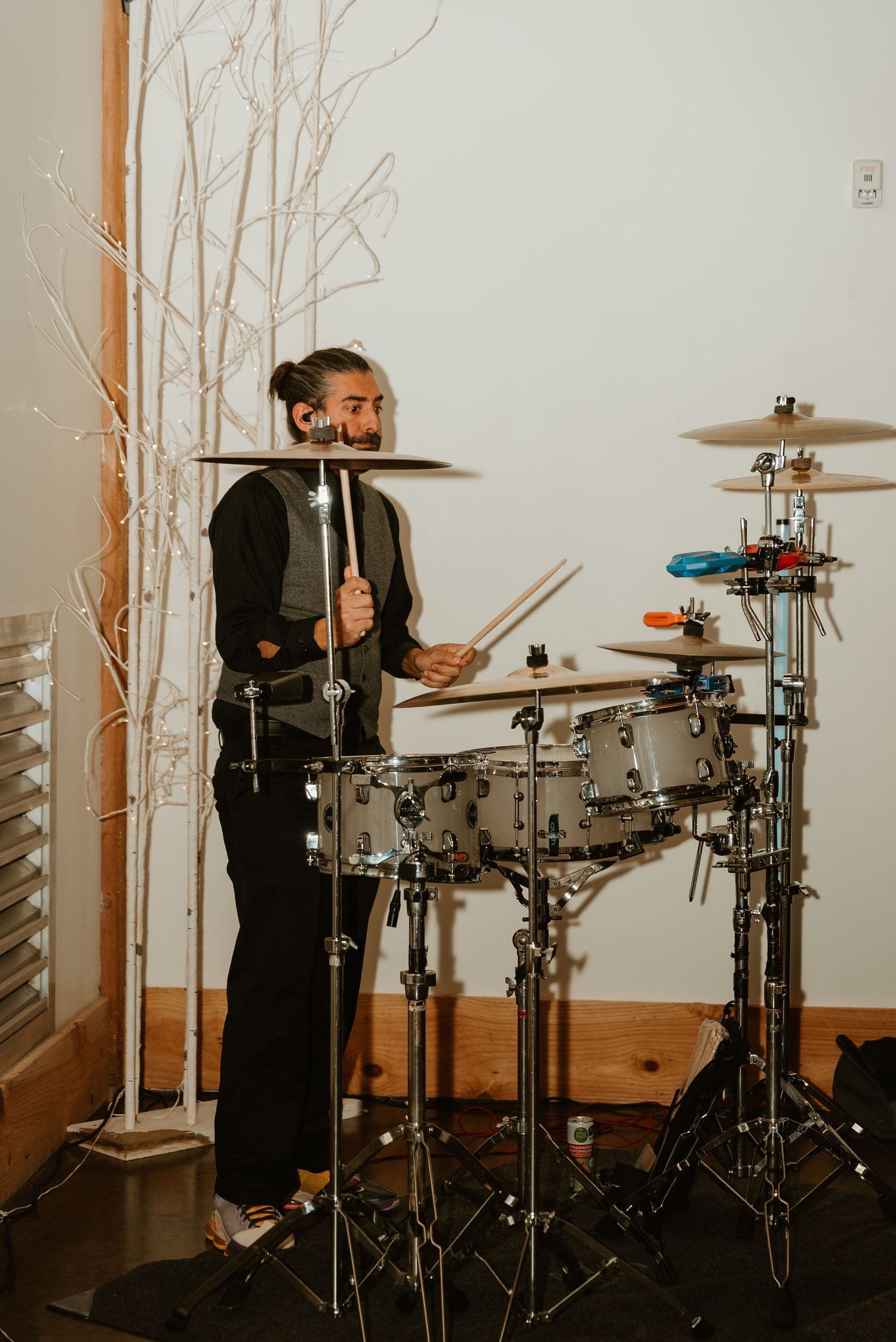 Smiling drummer with bun hairstyle playing drum kit beside white decorative branches indoors.