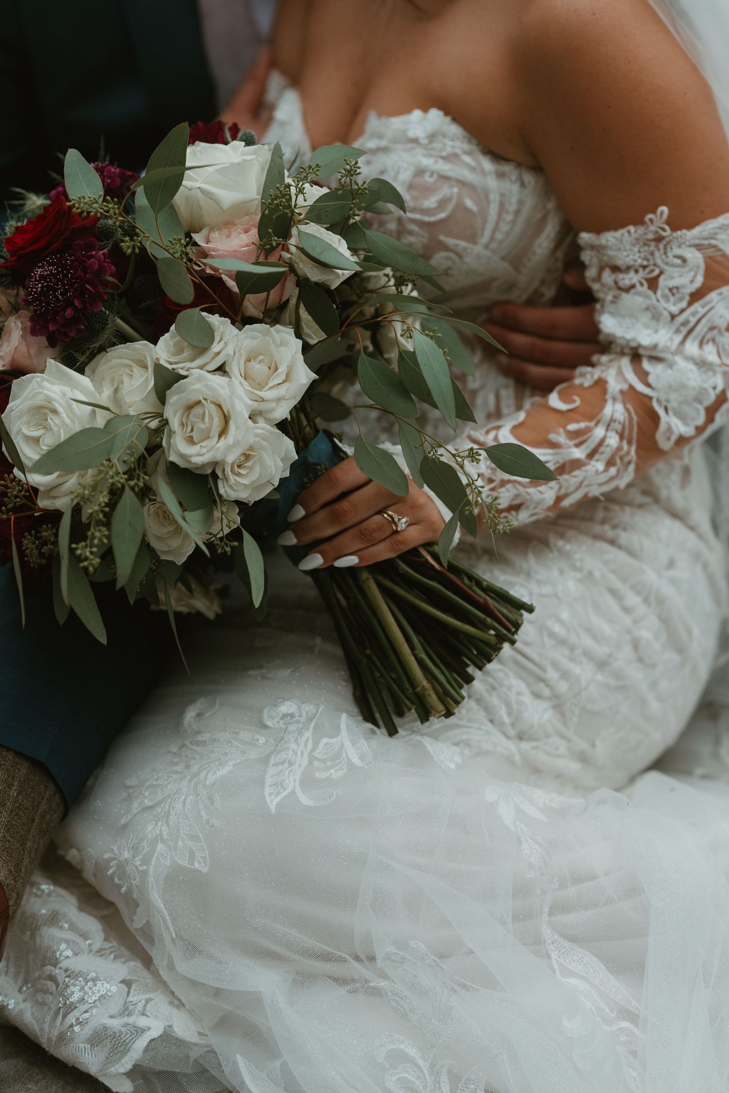 Close-up of bride holding elegant bouquet of white roses and greenery in lace off-shoulder wedding gown.